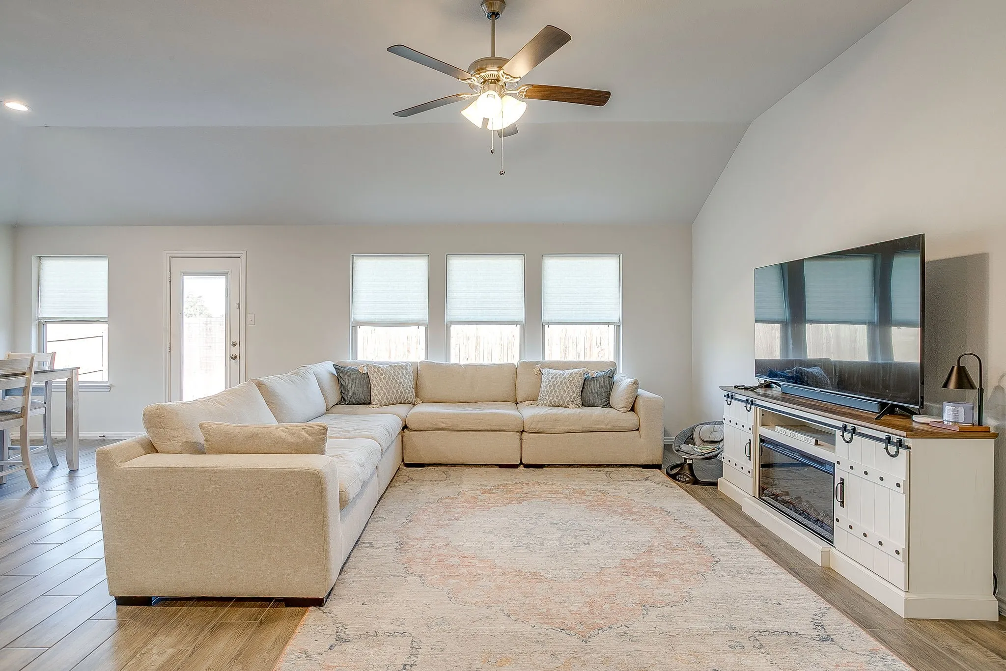 Vaulted ceiling with ceiling fan, wood-finish flooring, multiple windows with blinds, built-in entertainment console with electric fireplace insert, and a glass-paneled exterior door