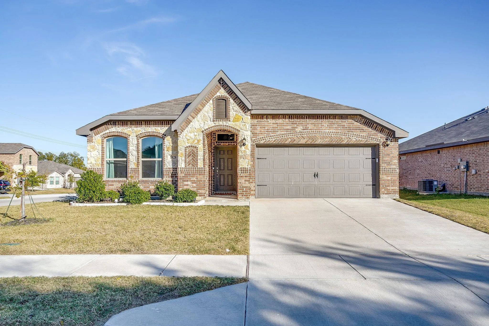 Brick and stone facade, arched entryway with a decorative door, attached two-car garage, concrete driveway, and a landscaped front yard