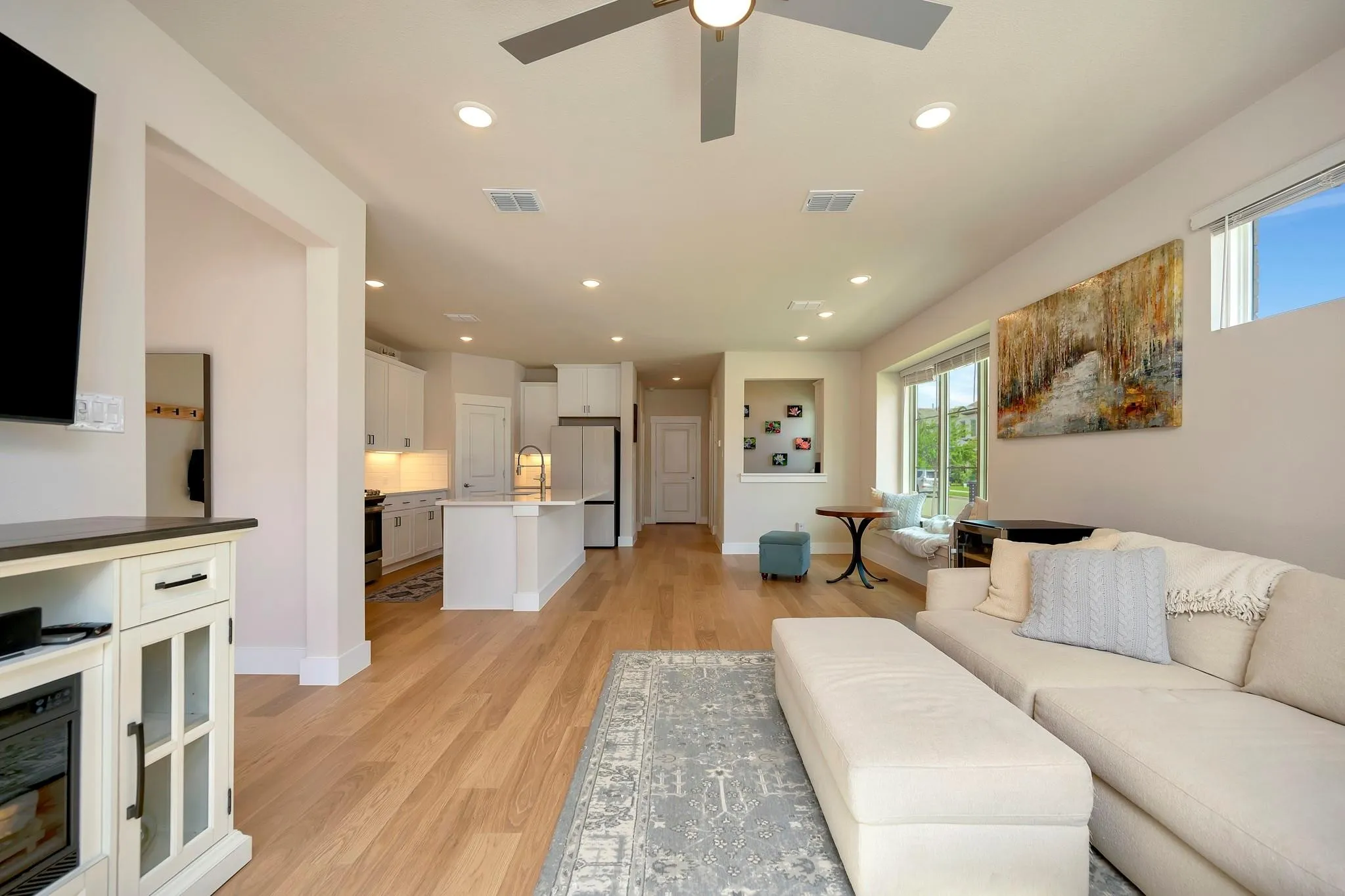 Wood-finish flooring throughout, recessed ceiling lighting, kitchen with white cabinetry and island, ceiling fan, and large windows