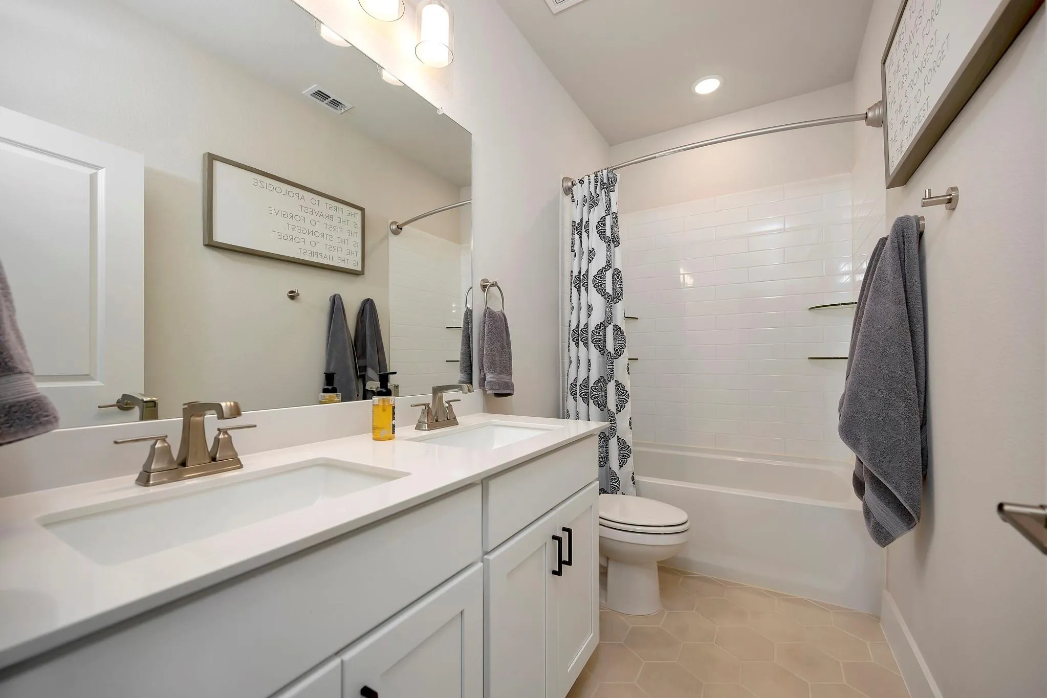 Dual vanity with integrated sinks, white cabinetry with dark hardware, a full-width mirror, a bathtub with subway tile surround, and hexagonal floor tiling