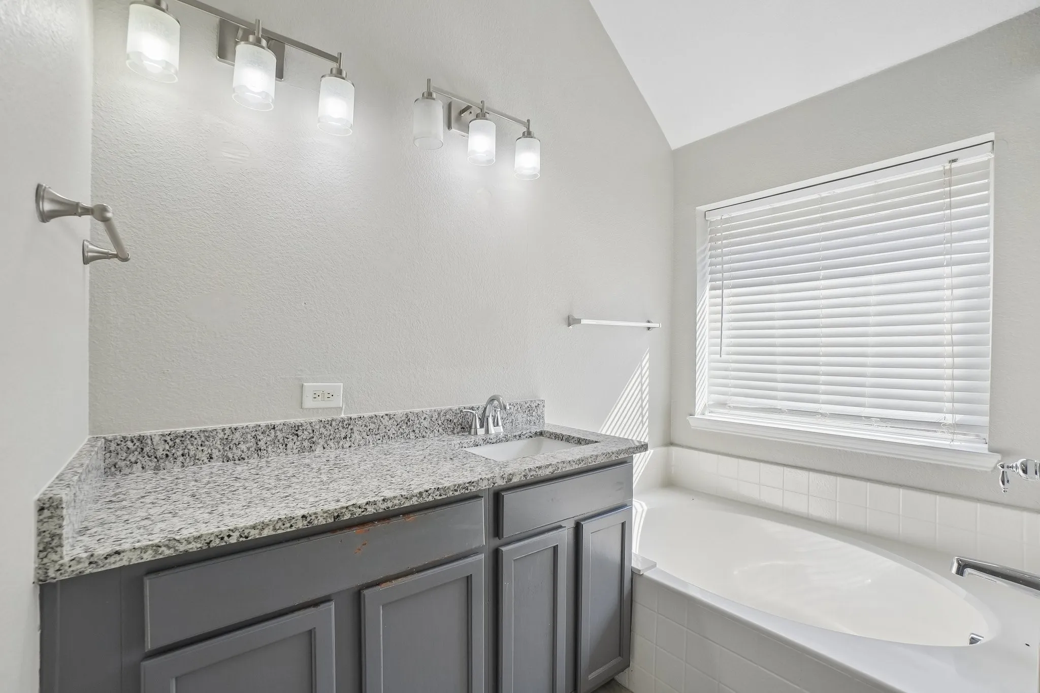 Dual vanity with gray cabinetry and granite countertops, single undermount sink, built-in bathtub with tiled surround, and a vaulted ceiling
