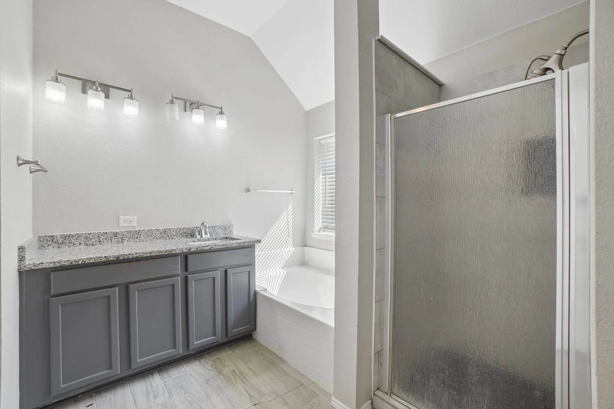 Dual vanity with gray cabinetry and speckled countertop, tiled flooring, built-in bathtub, and an enclosed shower with a textured glass door