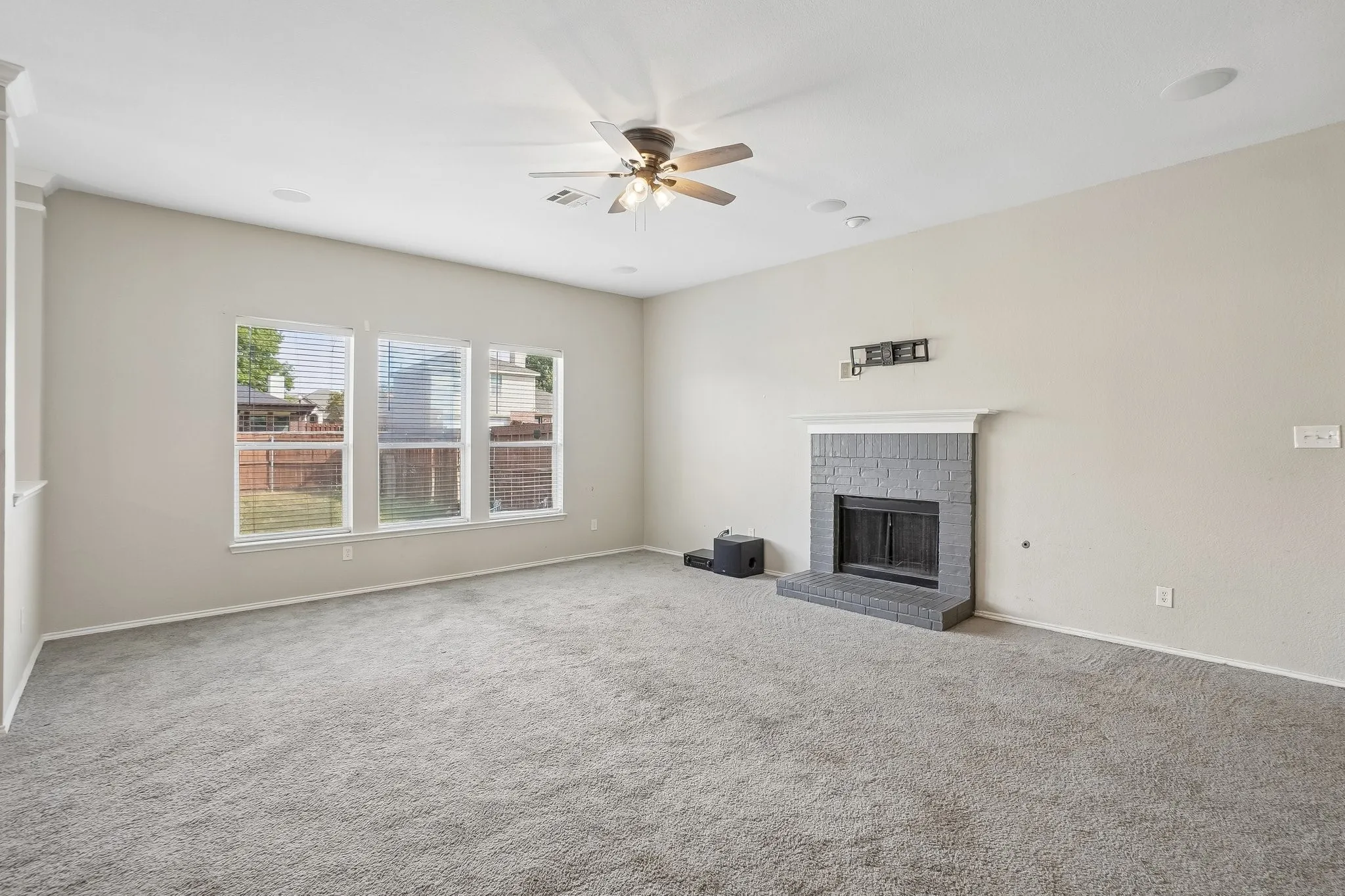 Spacious room featuring light-toned walls, a textured gray fireplace with a white mantel, three large windows, a ceiling fan, and recessed lighting