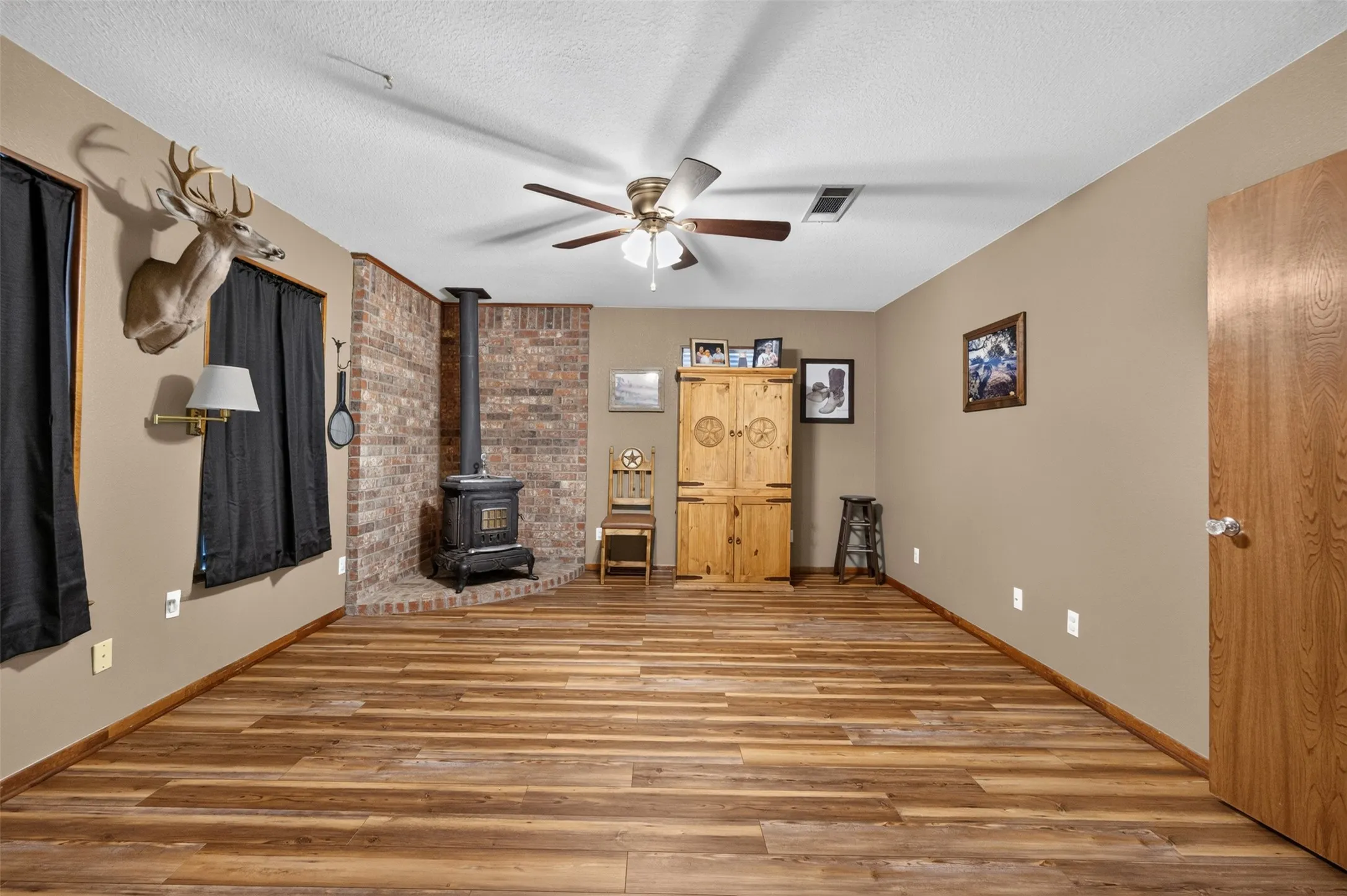 Wood-finish flooring throughout, brick accent wall, wood-burning stove, and ceiling fan