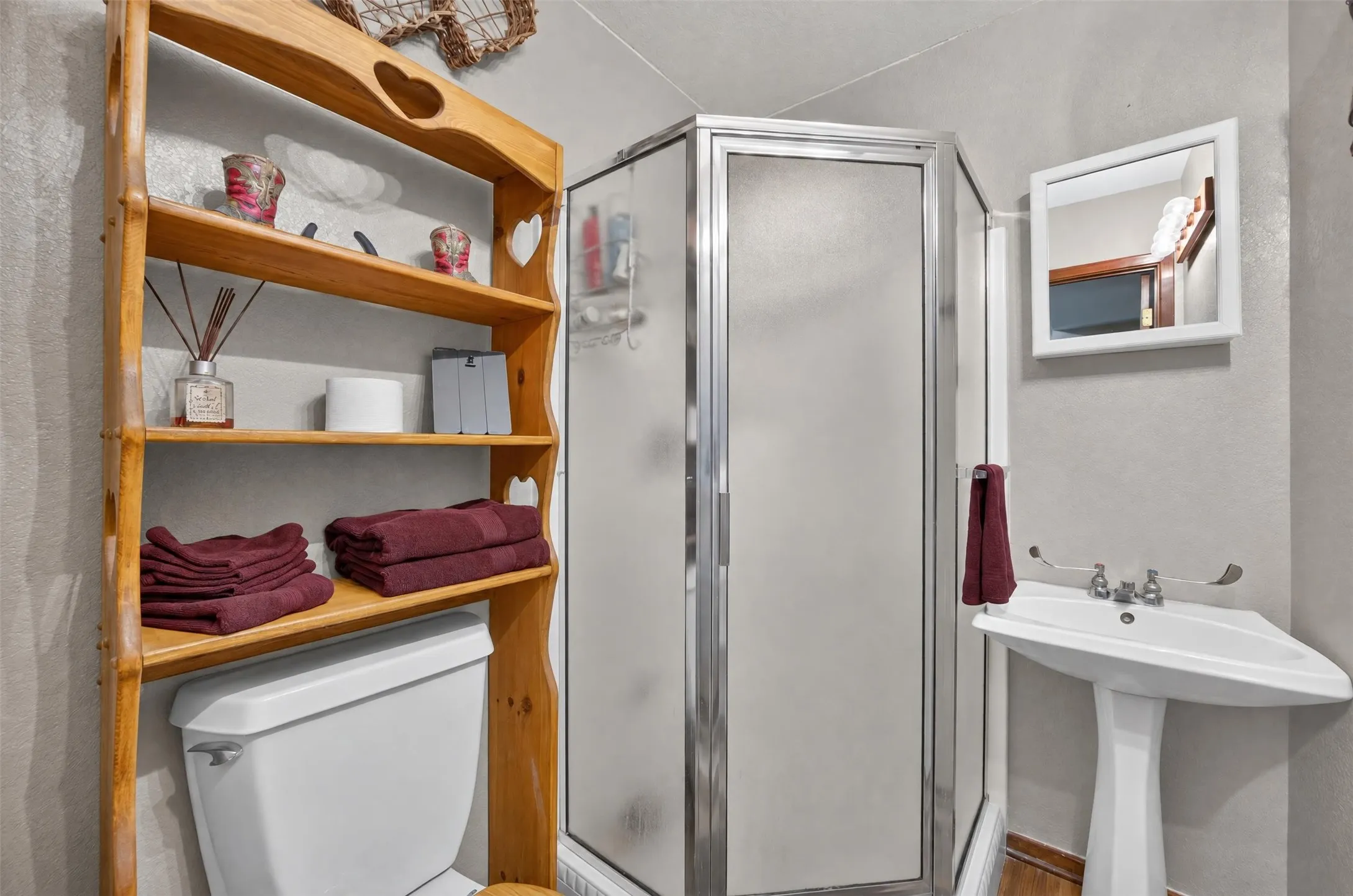 Corner shower with frosted glass enclosure, pedestal sink, wall-mounted mirror, and wood shelving unit