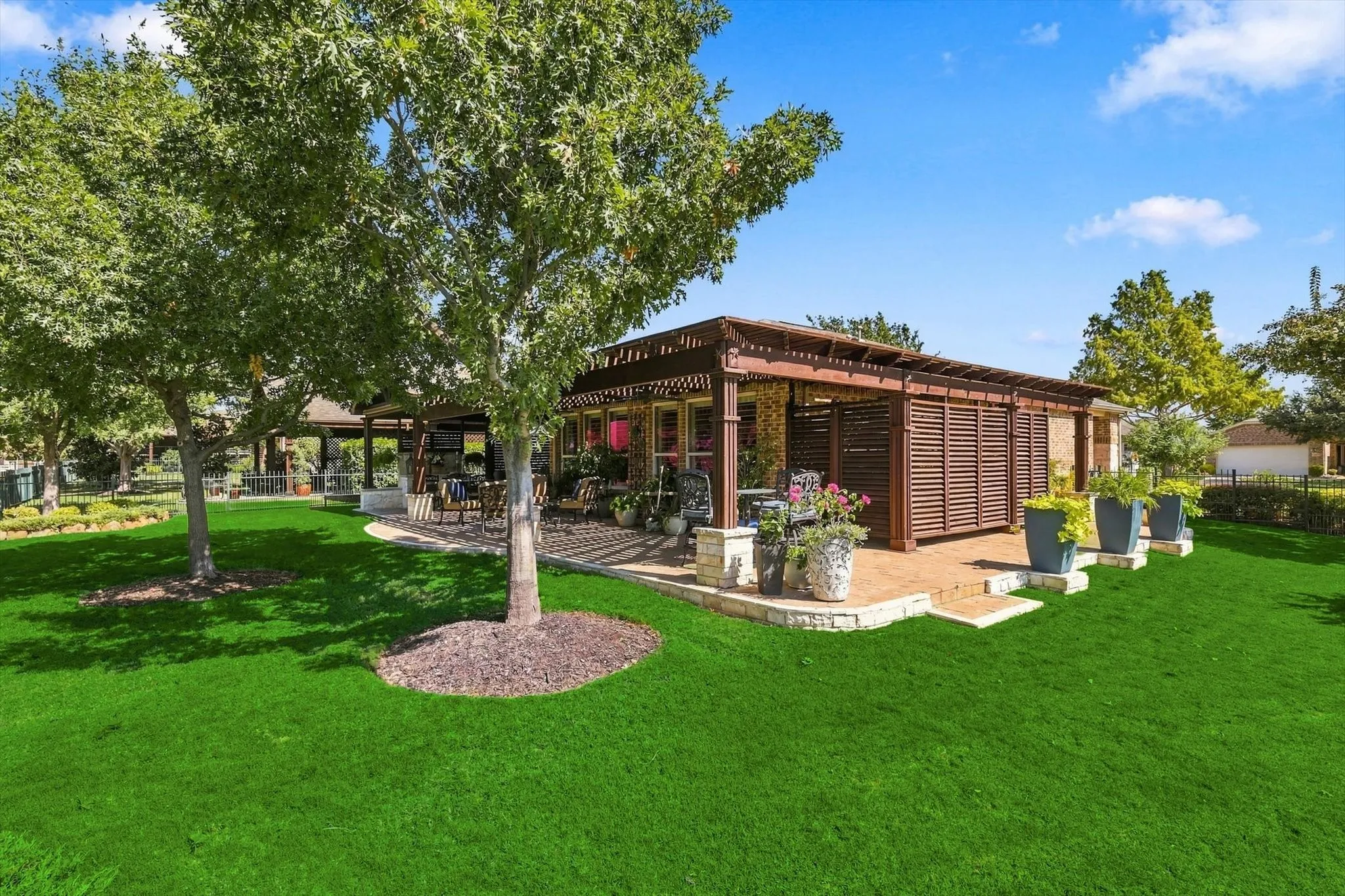 Green lawn with mature trees, a multi-level stone patio, an extended large wooden pergola, and slatted privacy screening for storage