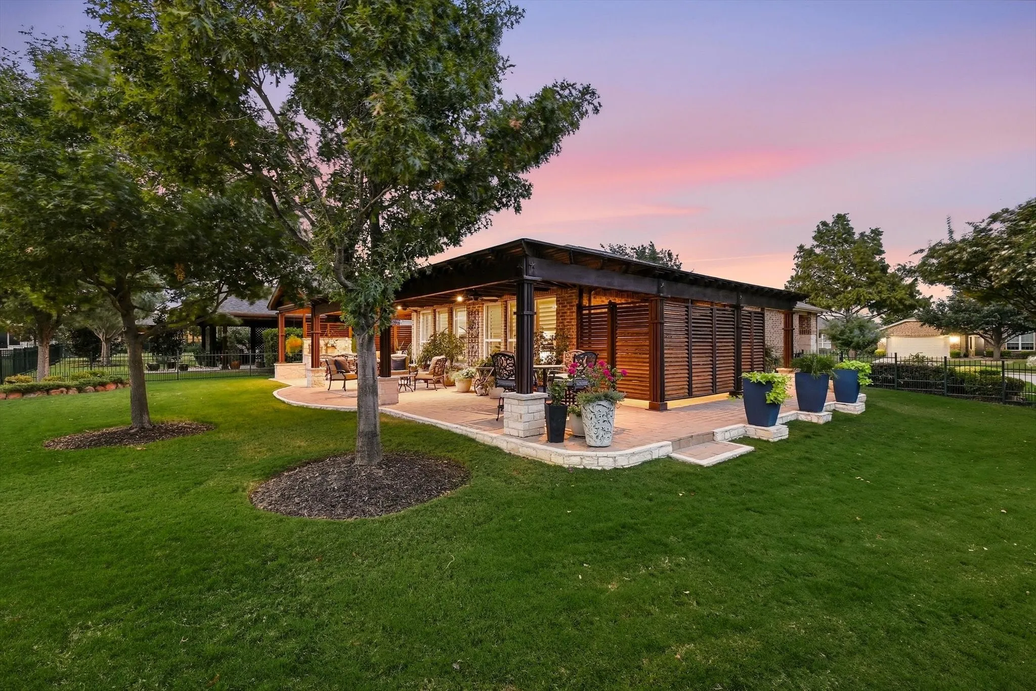 Expansive lawn space featuring mature trees, a multi-level patio with stone retaining walls, and a covered outdoor living area with wood slat privacy screens