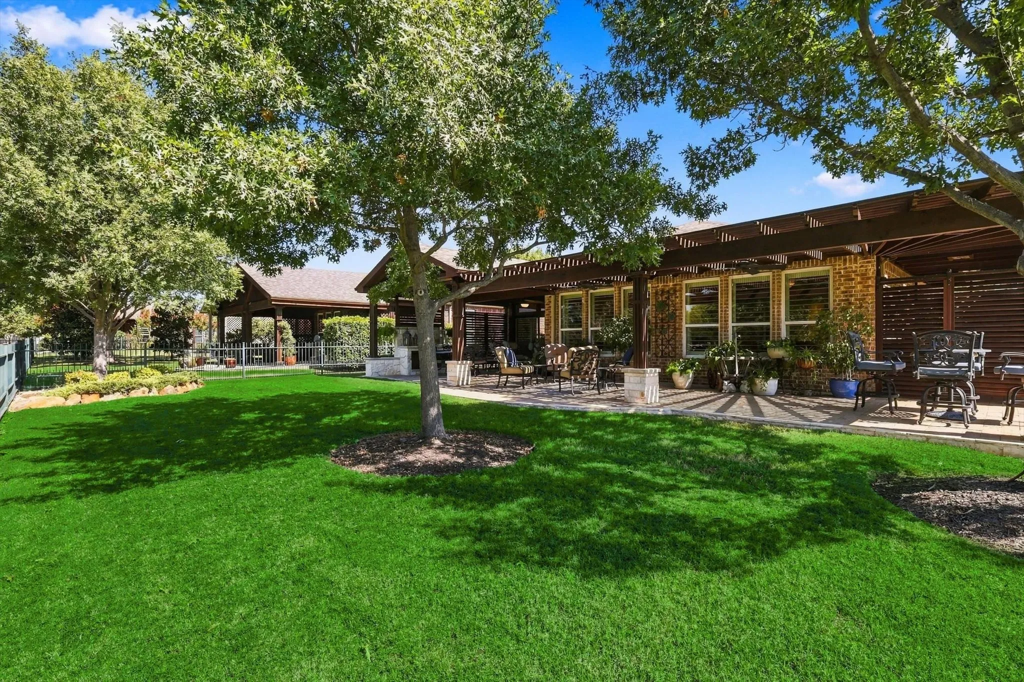 Expansive green lawn, mature trees providing natural shade, paver stone patio with a wood pergola, brick exterior, and multiple large windows
