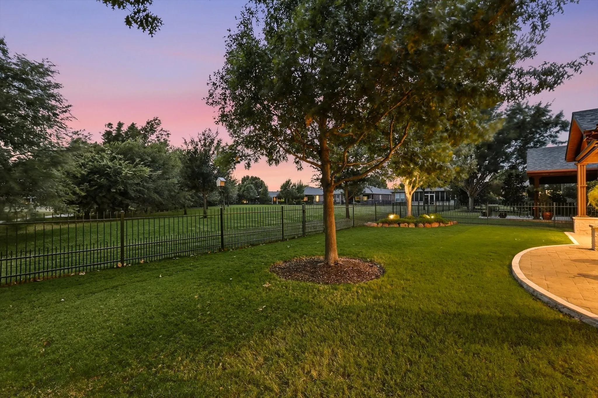 Expansive lawn with mature trees, a black metal fence, a curved paver patio, and recessed landscape lighting