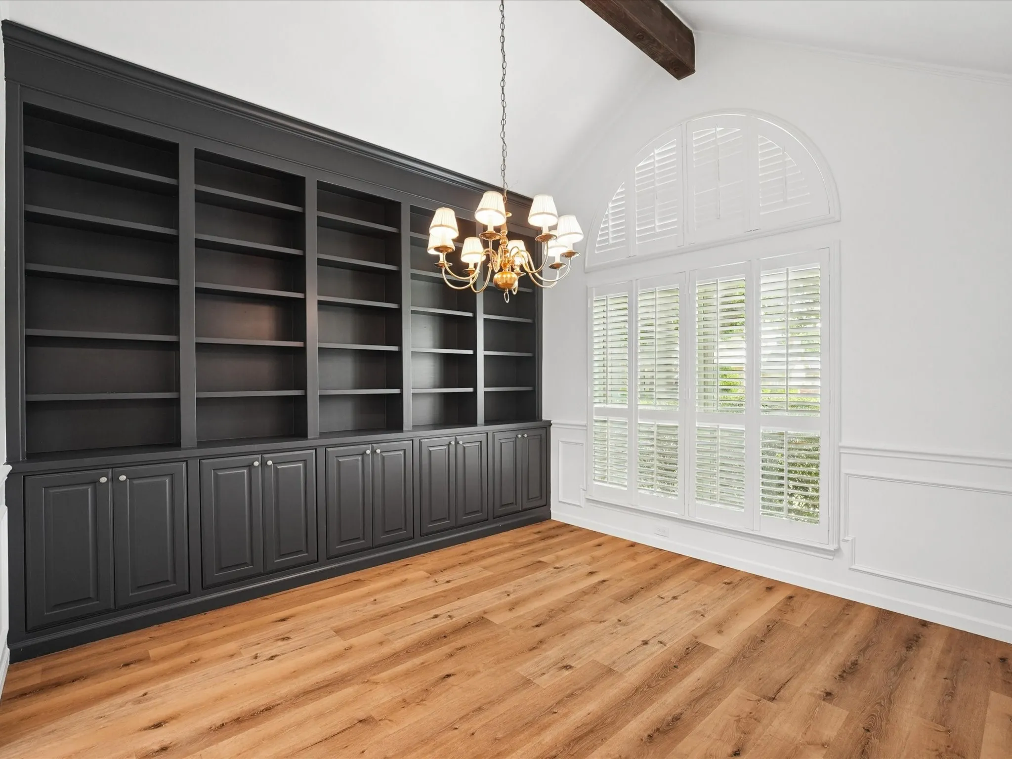 Expansive built-in shelving with cabinetry, wood-finish flooring, vaulted ceiling with exposed beam, arched window with plantation shutters, and decorative wall molding