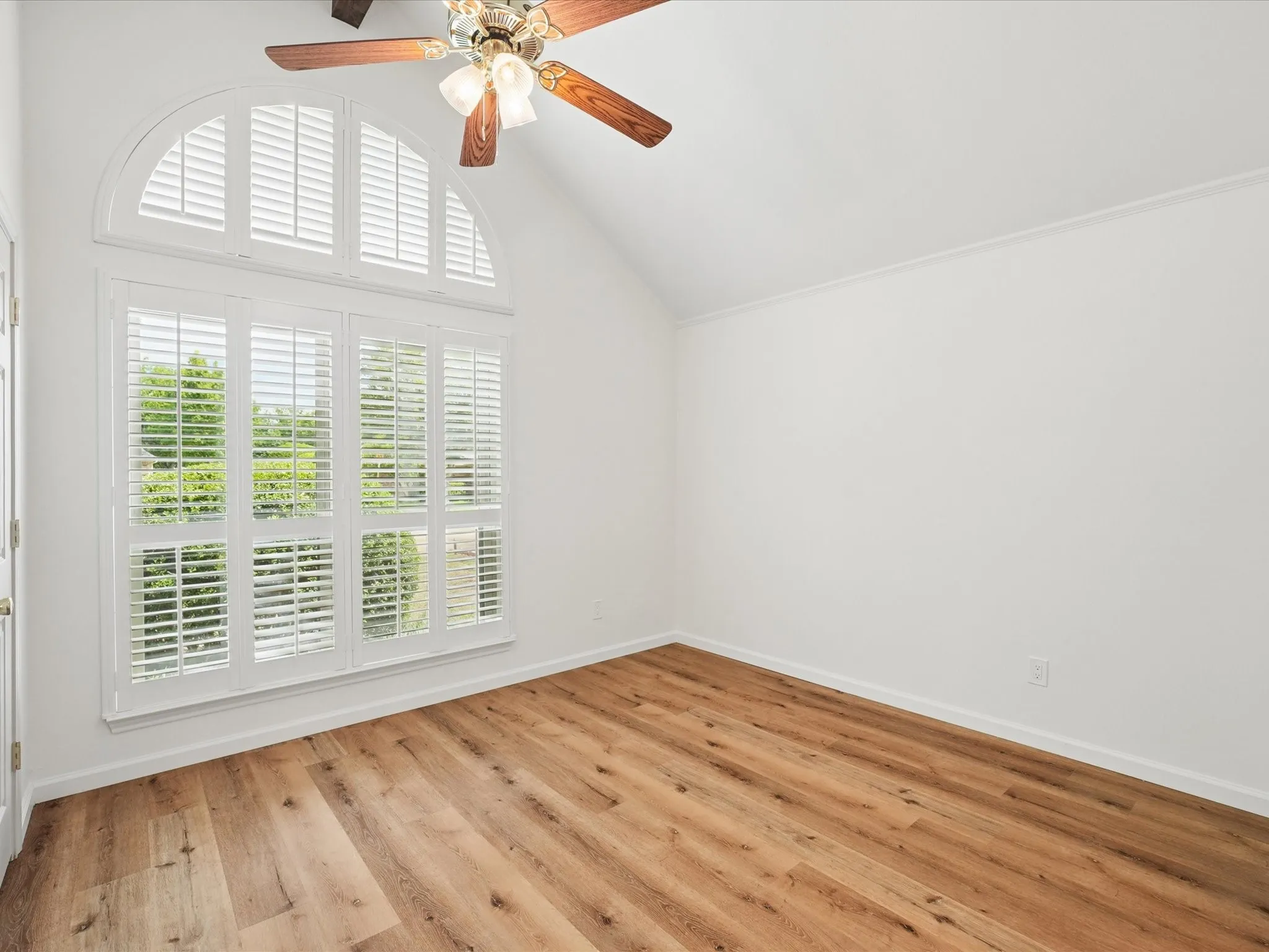 Wood-finish flooring, vaulted ceiling, arched window, white plantation shutters, and ceiling fan with lighting fixture