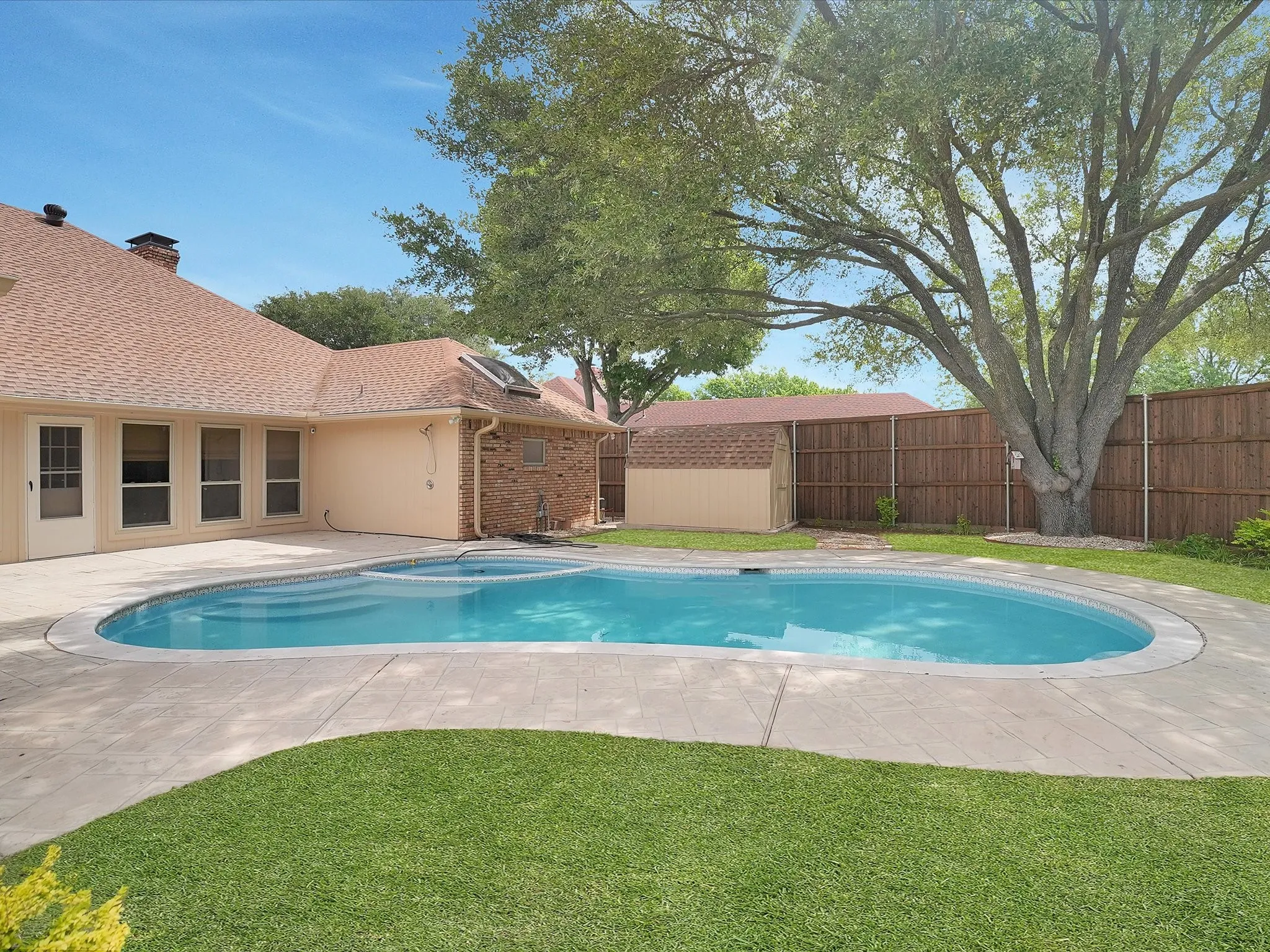 In-ground swimming pool with integrated spa, concrete decking, mature tree, manicured lawn, and privacy fence