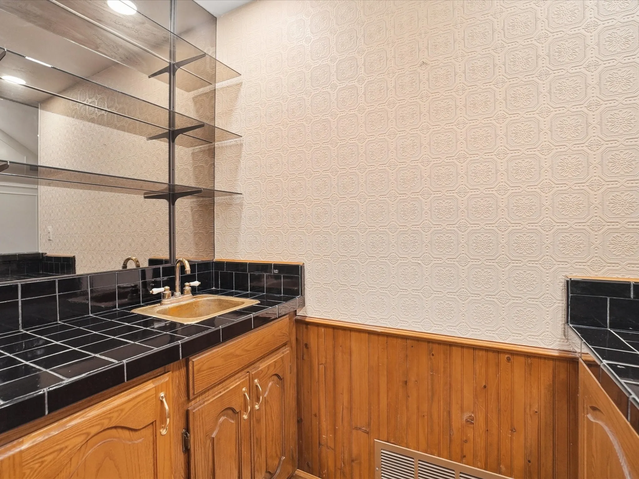 Built-in wet bar featuring a brass-tone sink, brushed nickel faucet, black tile countertop, wood cabinetry, and a mirrored backsplash with glass shelving