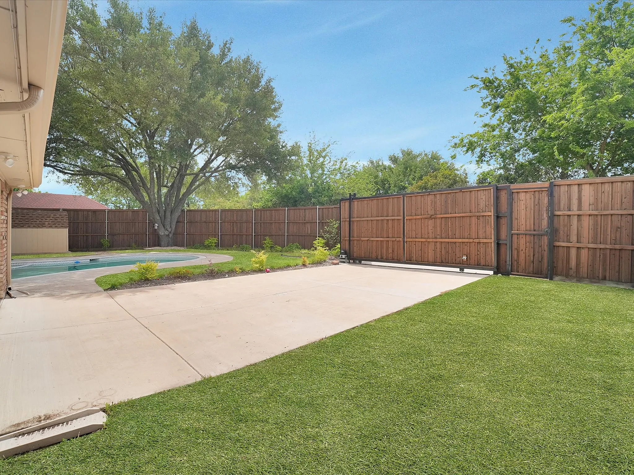Expansive backyard featuring a concrete patio, a swimming pool, a large mature tree, a privacy fence, and a sliding gate
