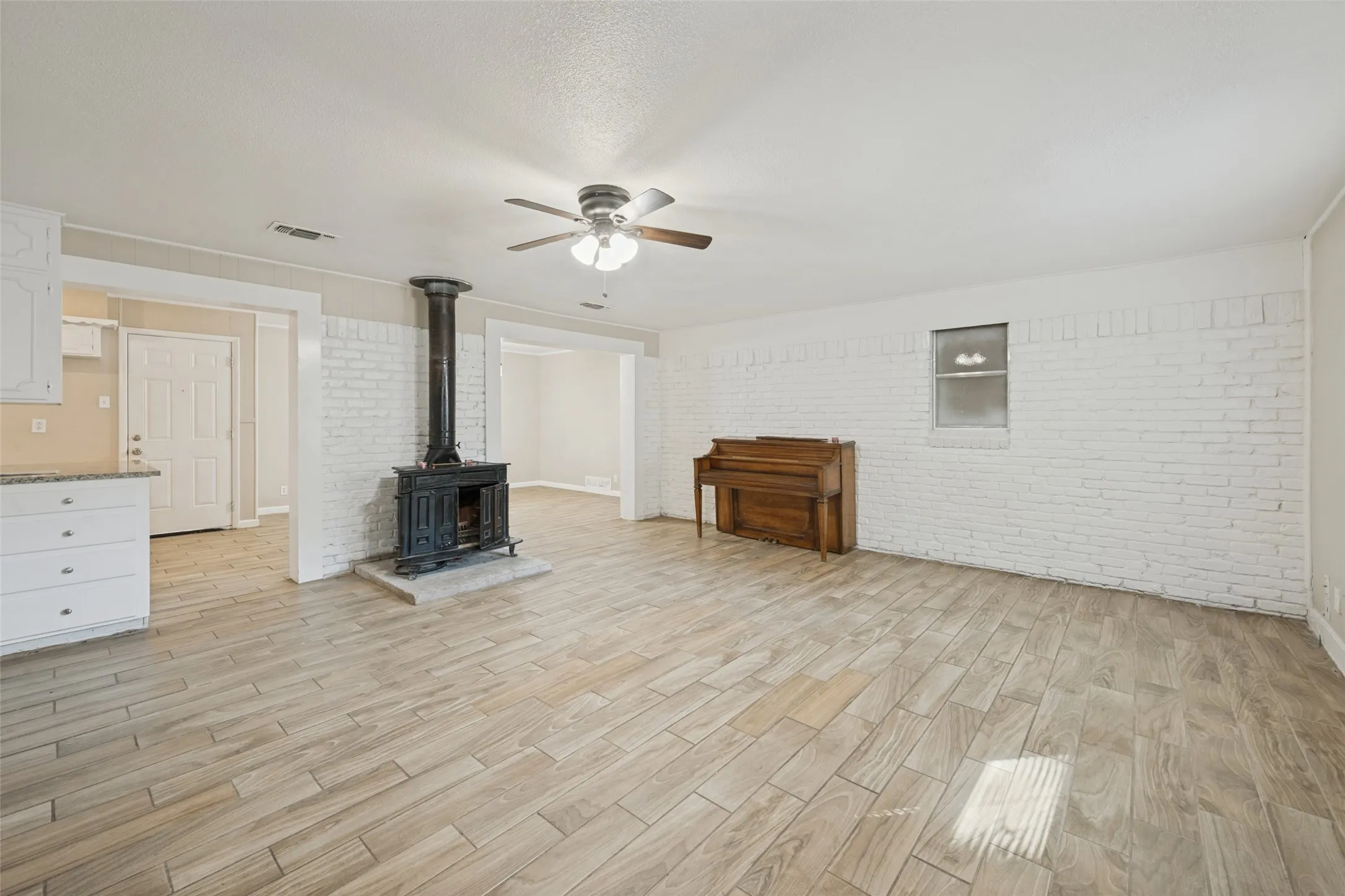 Wood-finish flooring, white brick accent wall, wood-burning stove, ceiling fan, and white cabinetry with granite-style countertops