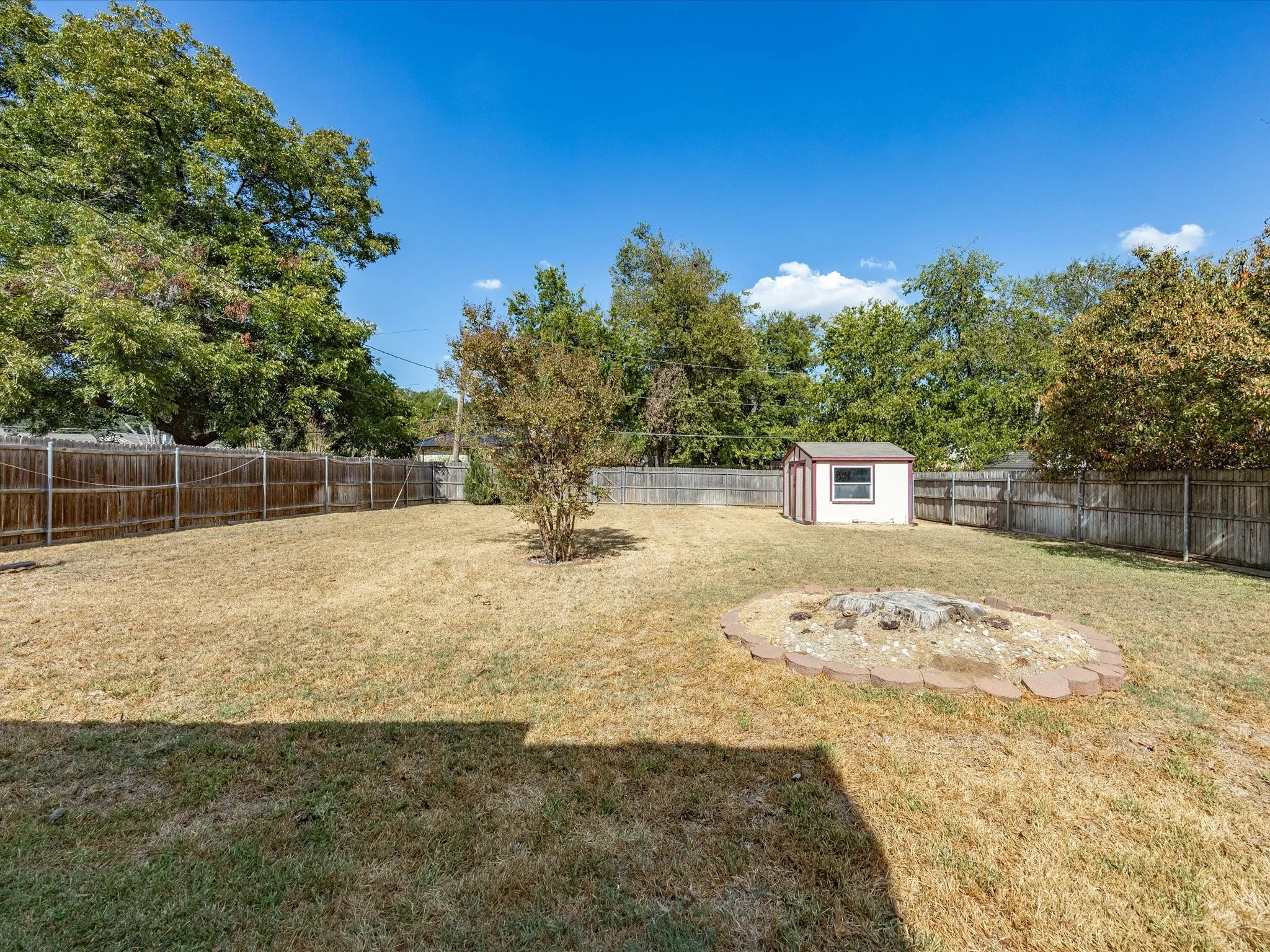 Expansive backyard featuring a wood fence, mature trees, a detached storage shed, and a circular hardscaped area