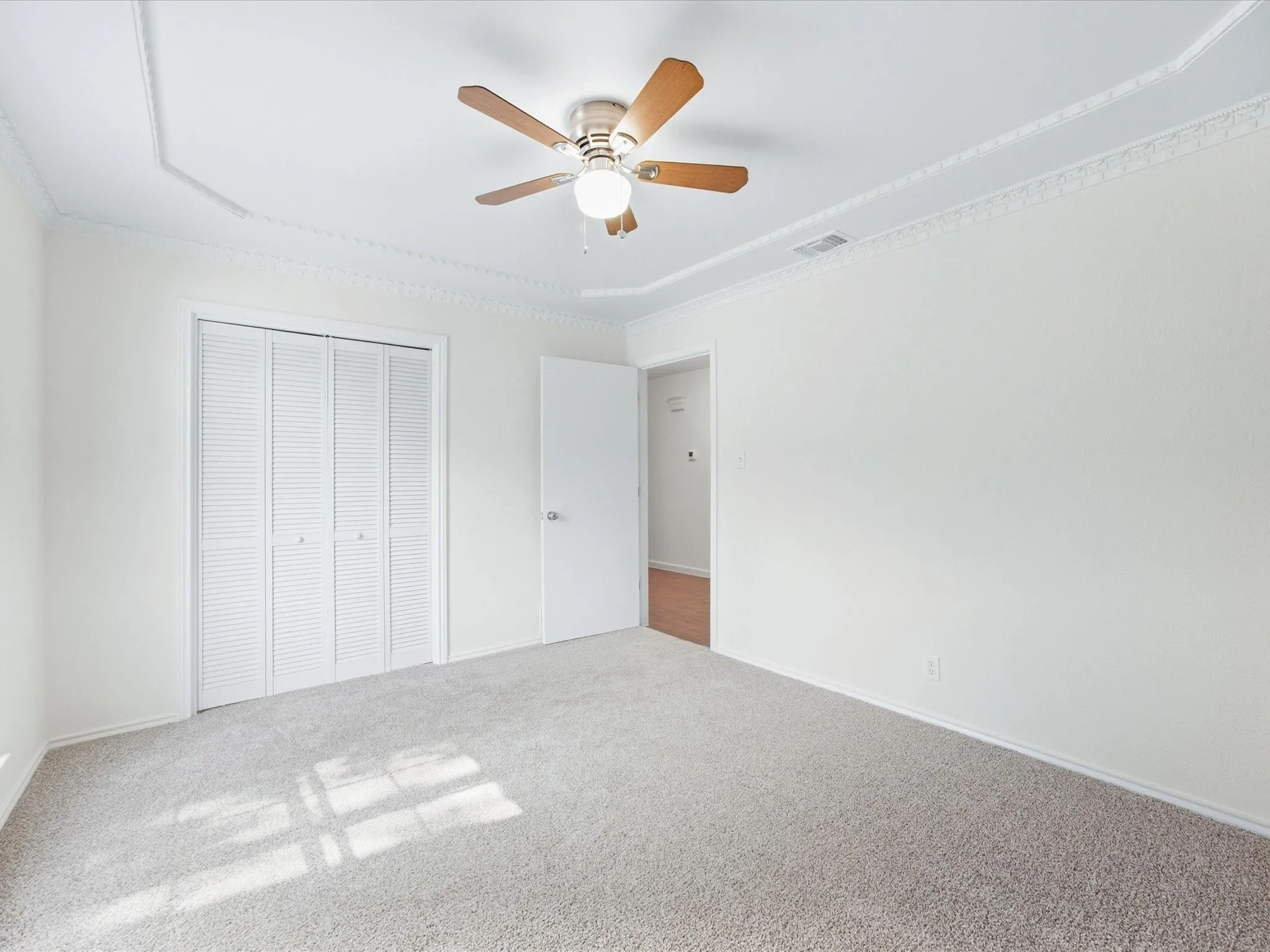 Neutral-toned carpeting, ceiling fan with light fixture, white louvered bifold closet doors, and crown molding accents