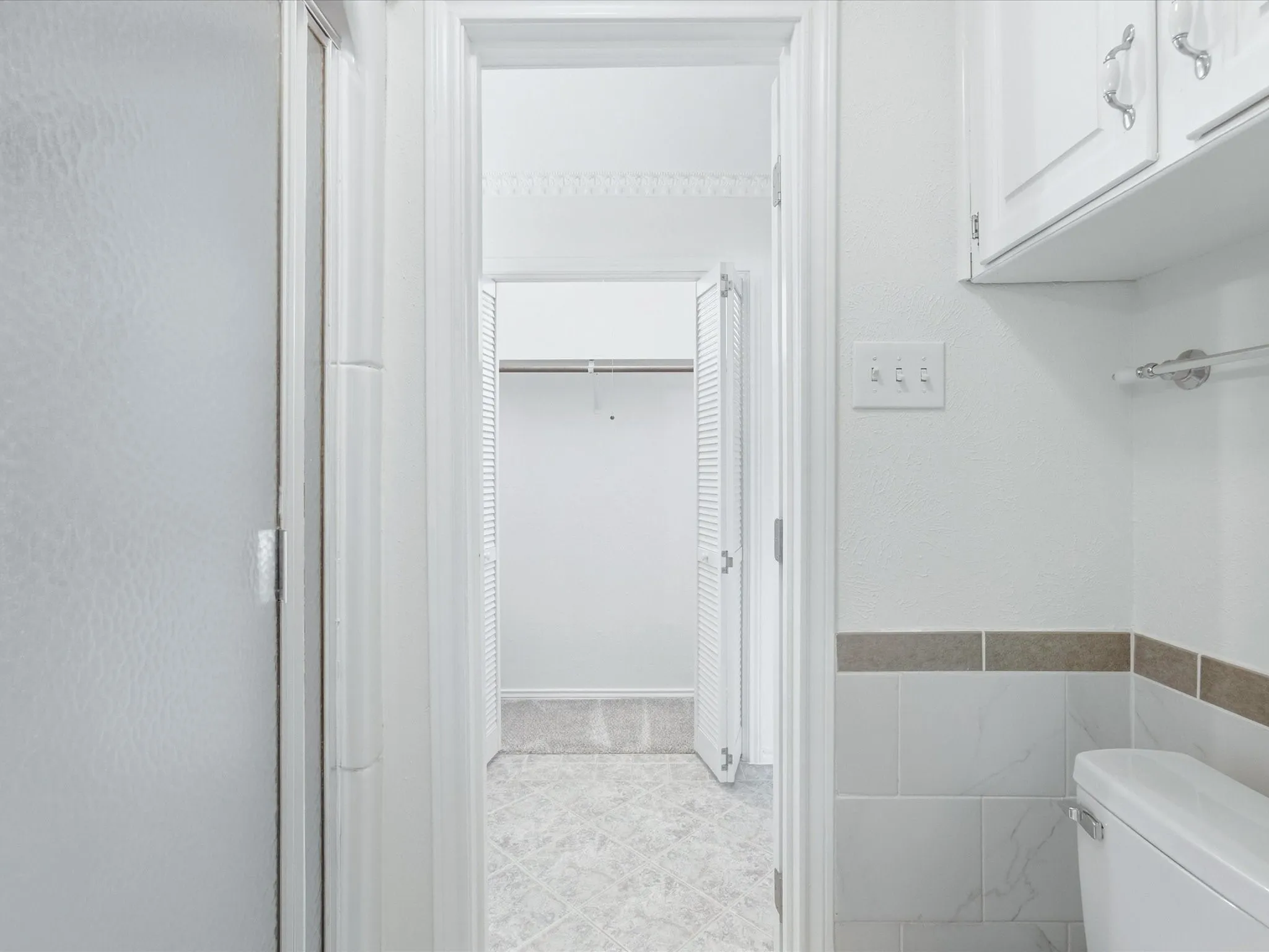 White shaker-style cabinetry with chrome hardware, white toilet, white and gray tiled wainscoting with a brown accent stripe, white textured wall finish, and white tile flooring