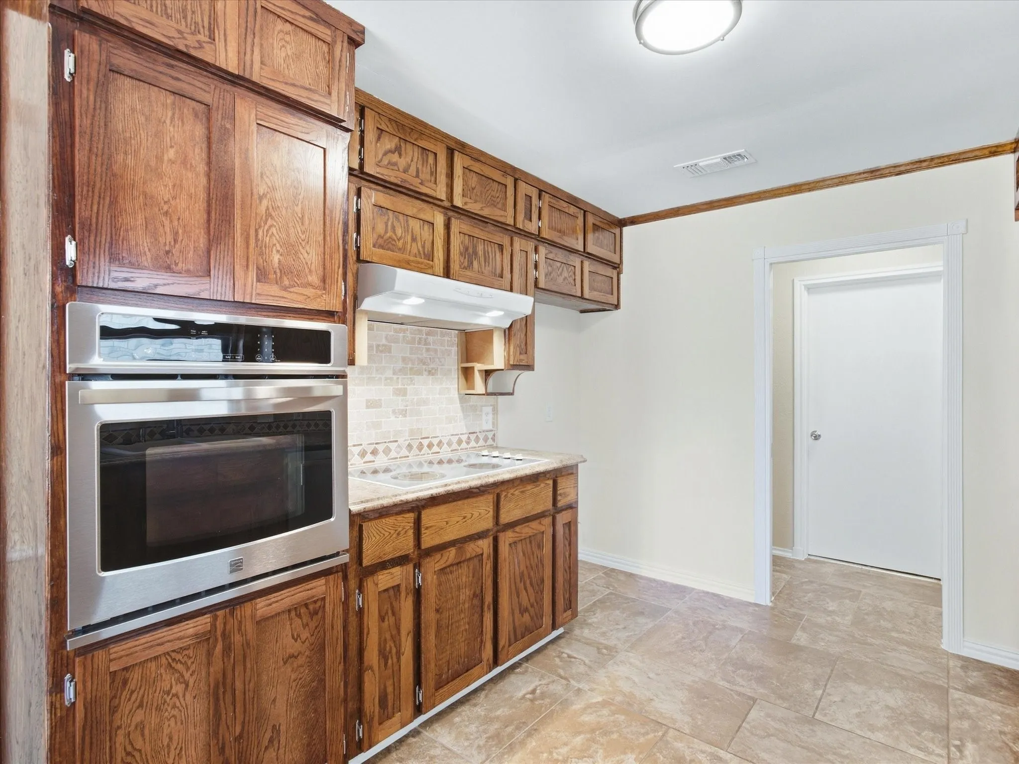 Stainless steel built-in oven, tile backsplash, wood cabinetry, stone-finish countertop, and tile flooring