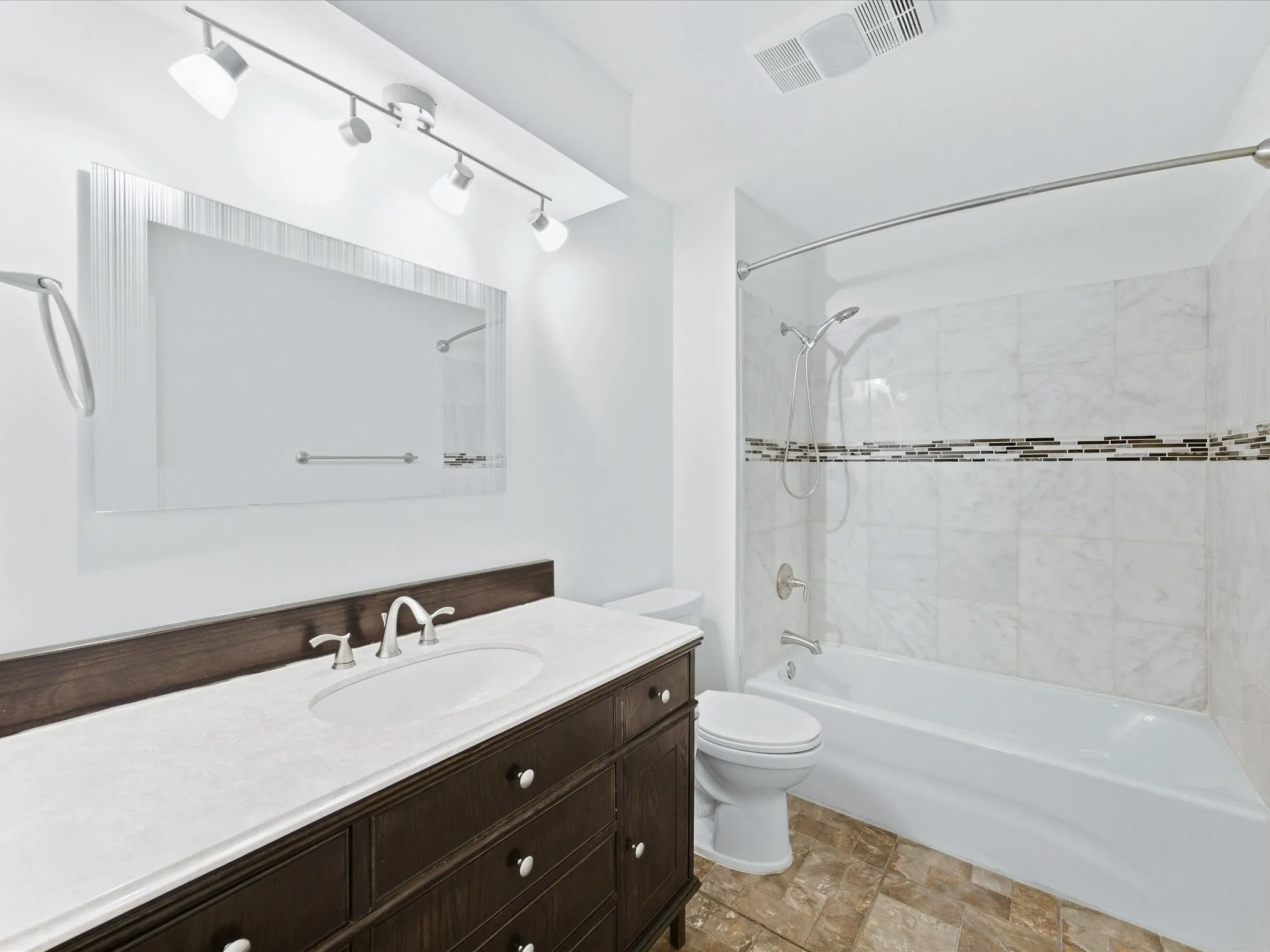 White tiled shower with decorative accent band, built-in bathtub, dark wood-finish vanity with countertop, and undermount sink with brushed nickel faucet