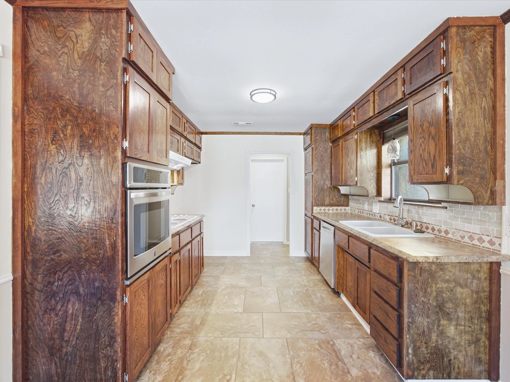 Galley kitchen featuring wood-finish cabinetry, tile flooring, stainless steel appliances, a tile backsplash, and a double basin sink