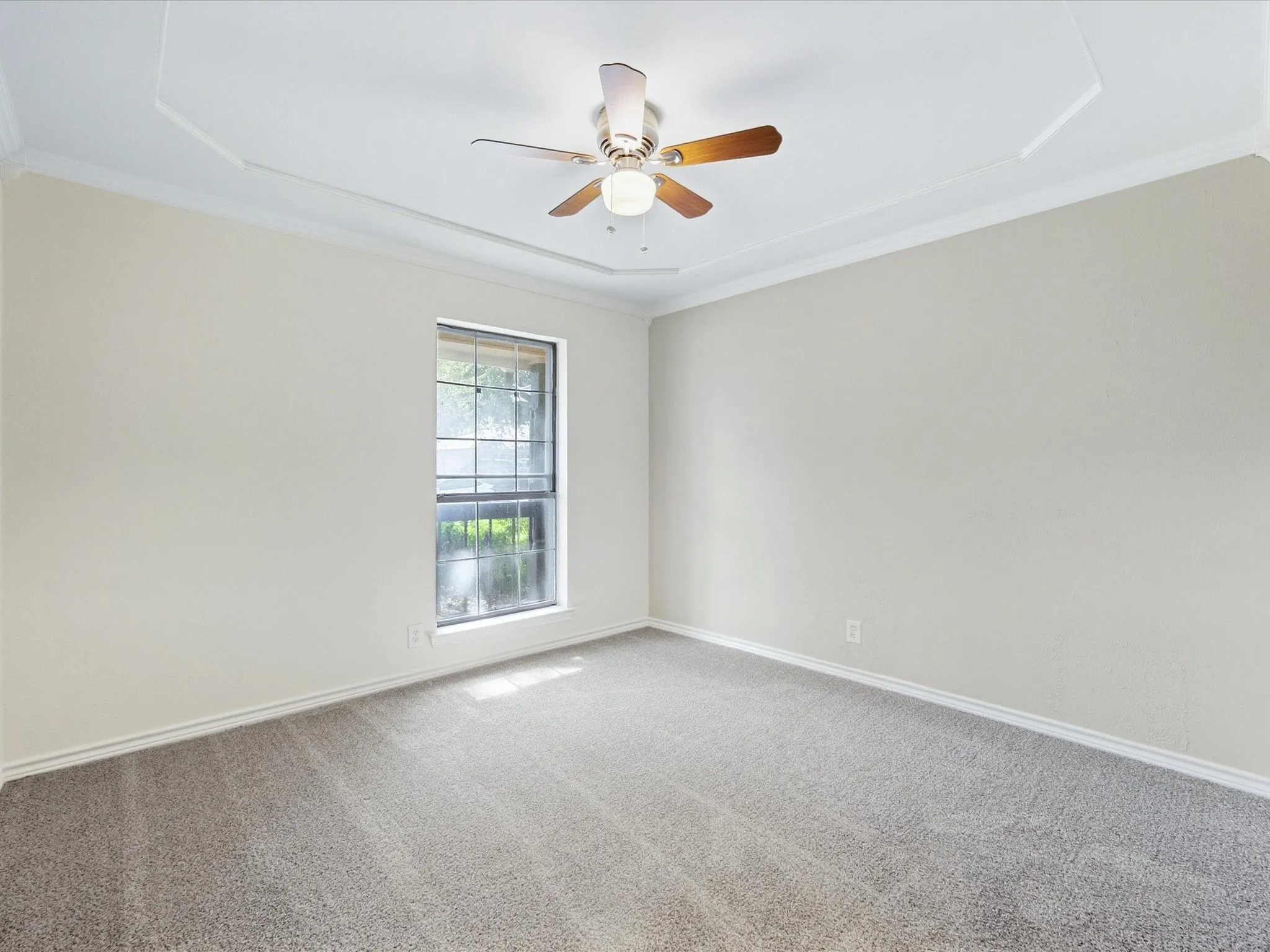 Tray ceiling with crown molding, ceiling fan with light fixture, window with grid pattern, and gray carpet flooring