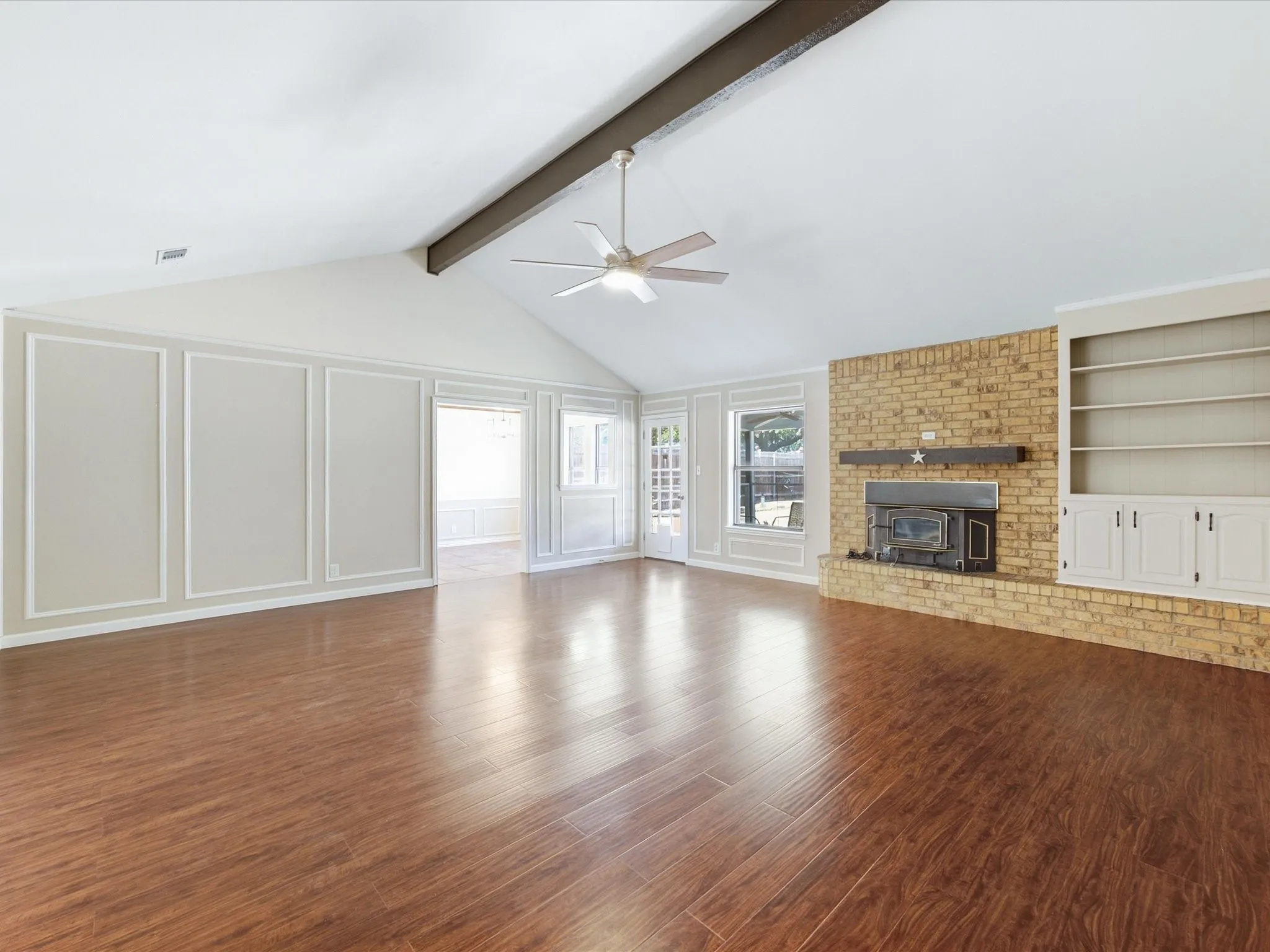 Wood-finish flooring, vaulted ceiling with exposed beam, brick fireplace with wood mantel, built-in shelving, and paneled wall detailing