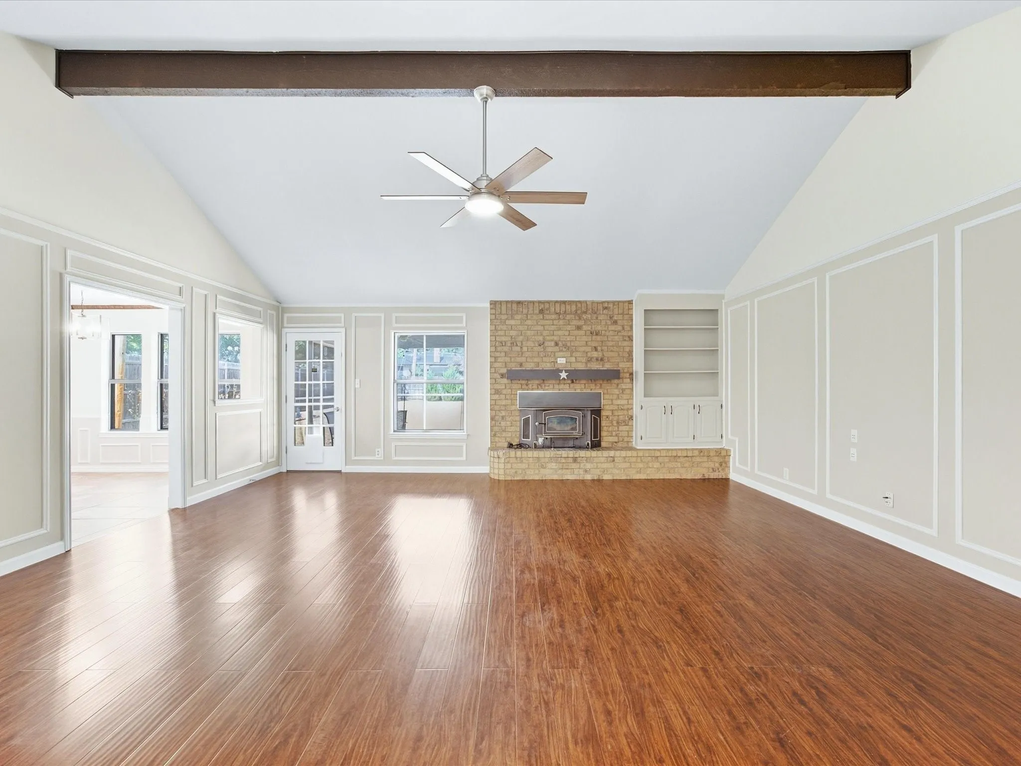Vaulted ceiling featuring a dark wood beam, wood-finish flooring, brick fireplace with wood stove insert, built-in shelving, and white paneled walls