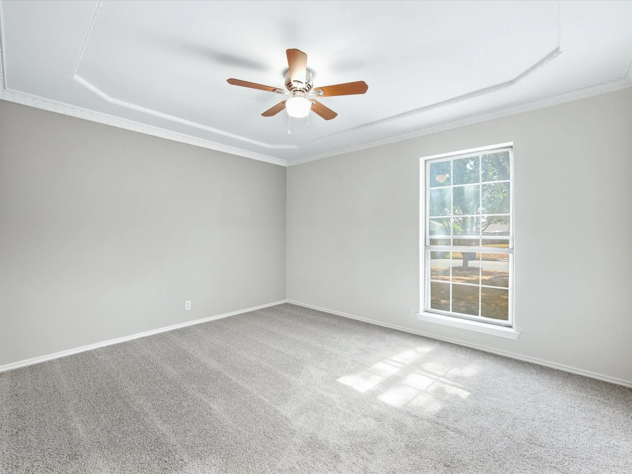 Recessed tray ceiling with crown molding, ceiling fan, large window, and light grey carpeting