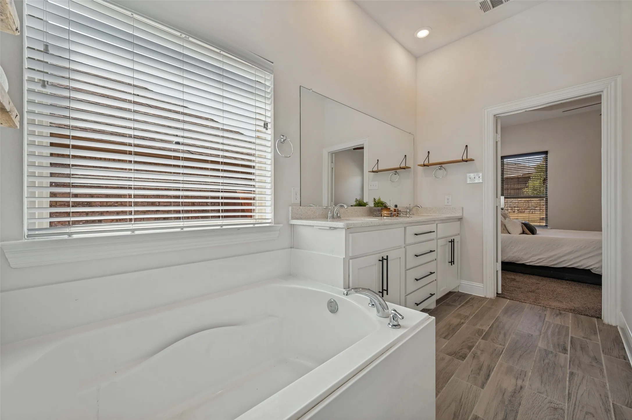 Soaking tub with a window, dual vanity with undermount sinks and quartz countertops, modern cabinetry with matte black hardware, and wood-finish flooring