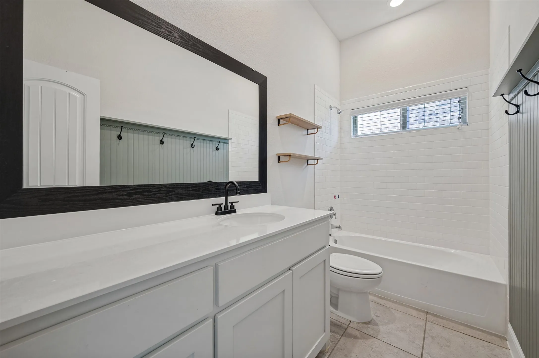 White subway tile bathtub surround, a window with blinds, a vanity with a white countertop, and a black-framed mirror