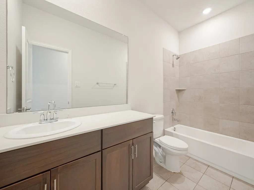 Vanity with white countertop and integrated sink, chrome faucet, wood-finish cabinetry, ceramic tile flooring, and tiled shower/tub combination with built-in niche