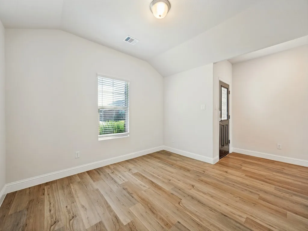 Wood-finish flooring, vaulted ceiling, single window with blinds, white walls, and a dark paneled door with frosted glass
