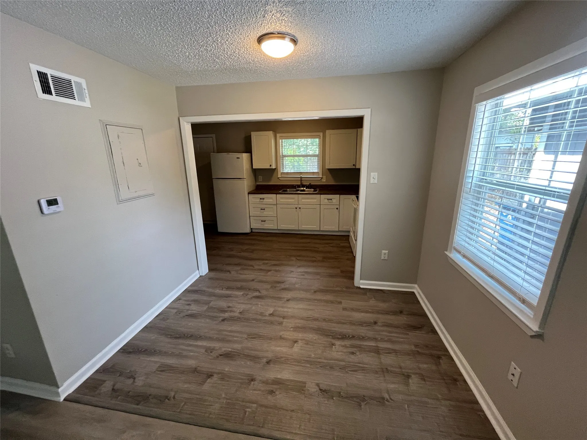Wood-finish flooring throughout, with a recessed kitchen featuring white cabinetry, a refrigerator, and a window with blinds
