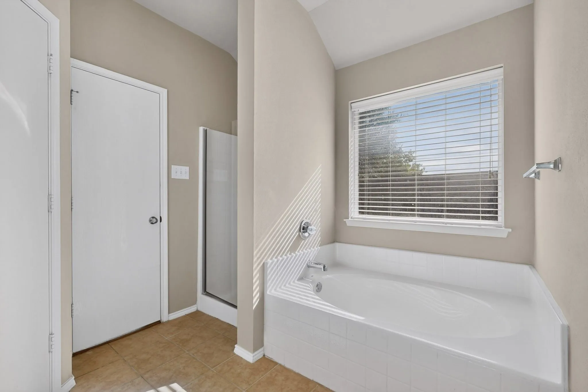 Built-in soaking tub with tile surround, separate glass-enclosed shower, large window with blinds, and neutral wall tones