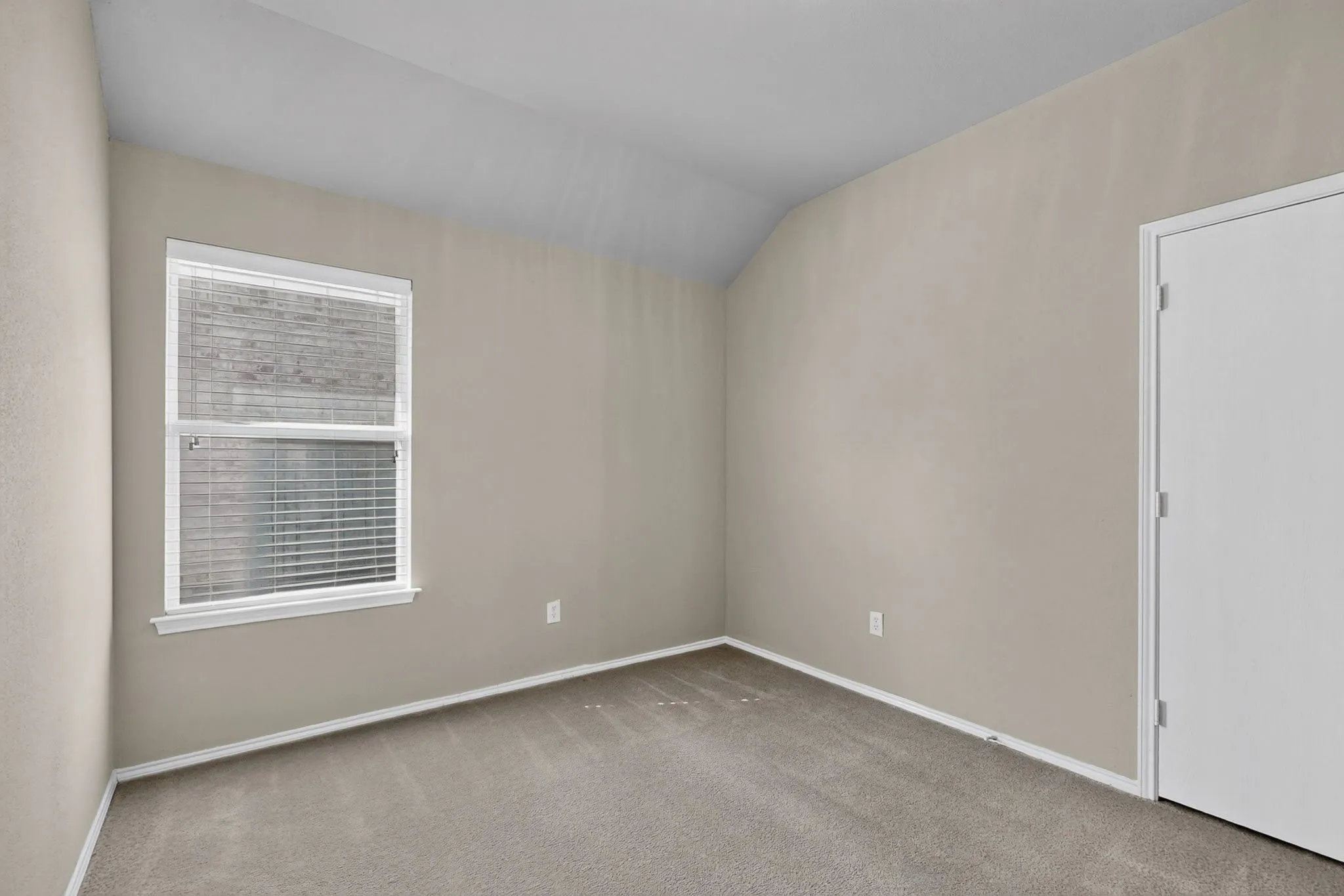 Neutral tone wall paint, vaulted ceiling, white trim window with blinds, light gray carpet, and white door