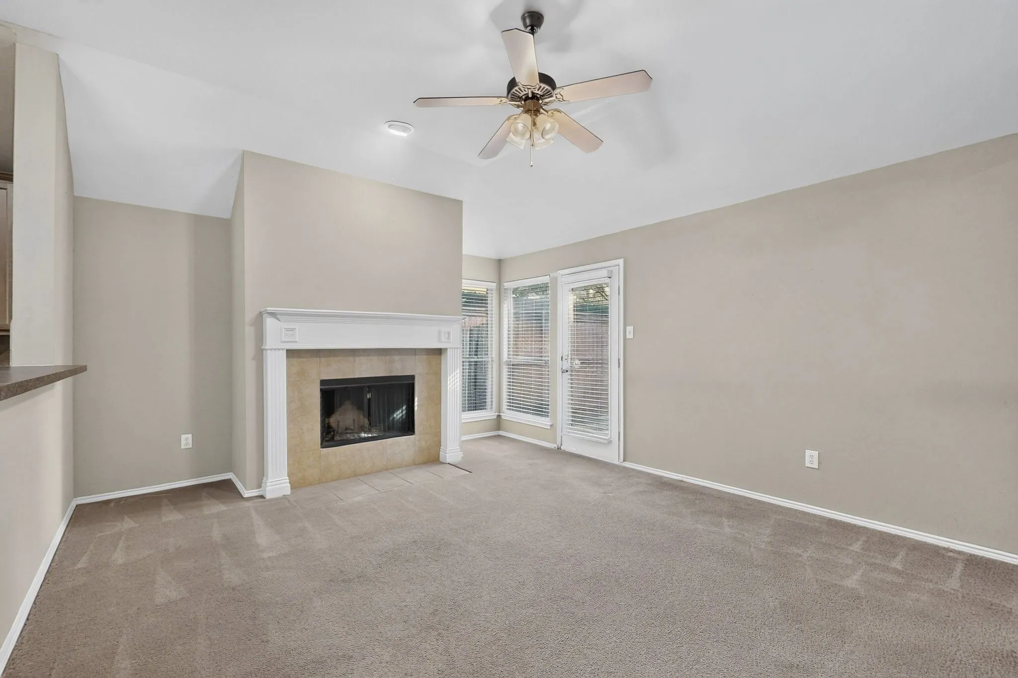 Carpeted living area featuring a fireplace with a white mantle, vaulted ceiling, ceiling fan, and access to an outdoor area through a glass door