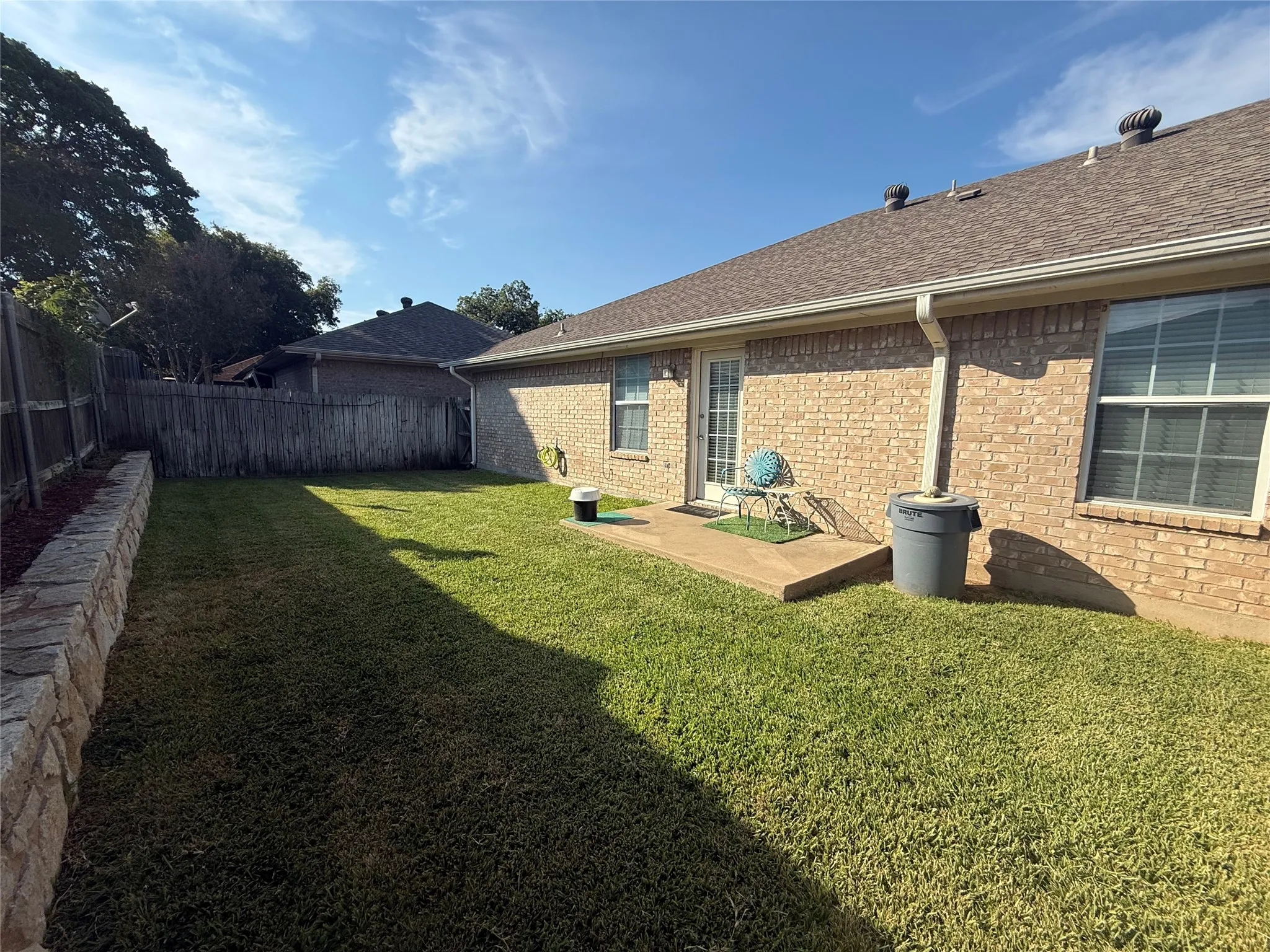 Brick exterior with shingle roofing, framed windows, a rear entry door, and a stone retaining wall
