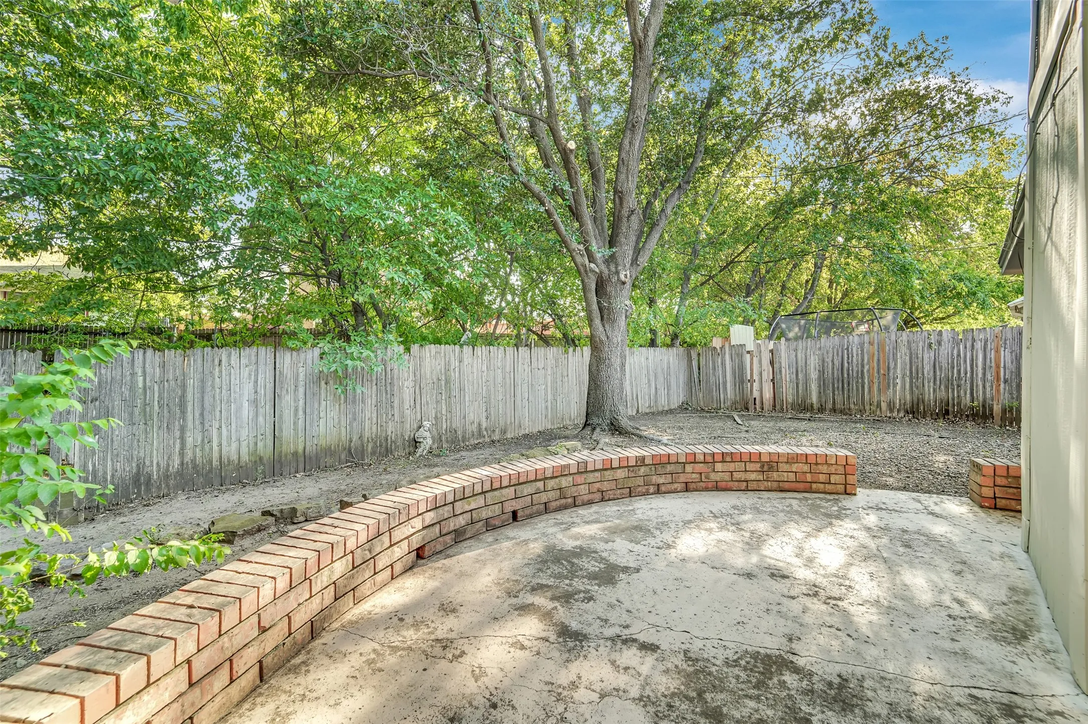 Expansive paved outdoor area with a curved brick planter, mature trees, and a wood fence enclosure