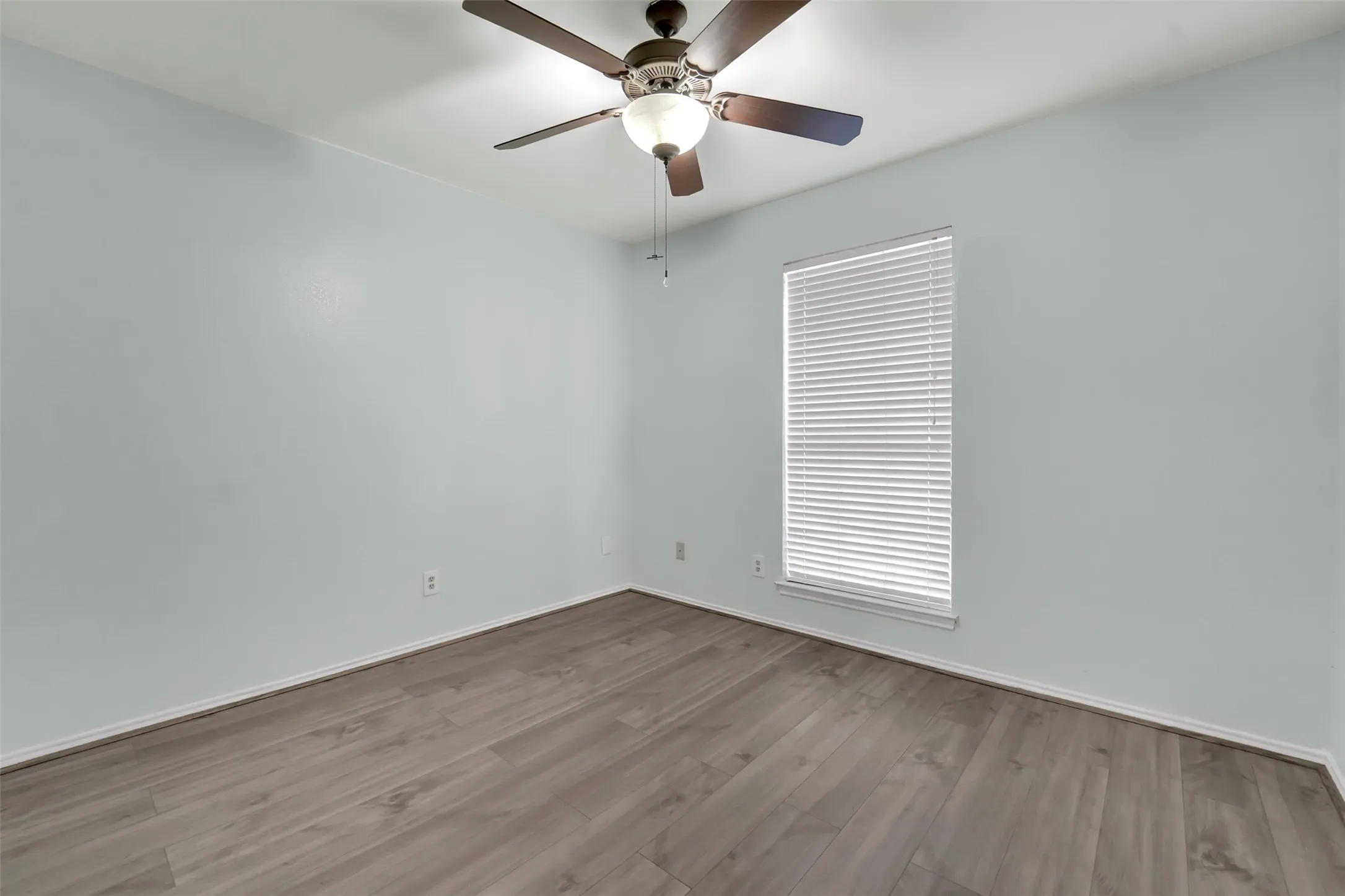 Wood-finish flooring with a neutral wall palette, featuring a window with blinds and a ceiling fan with integrated lighting