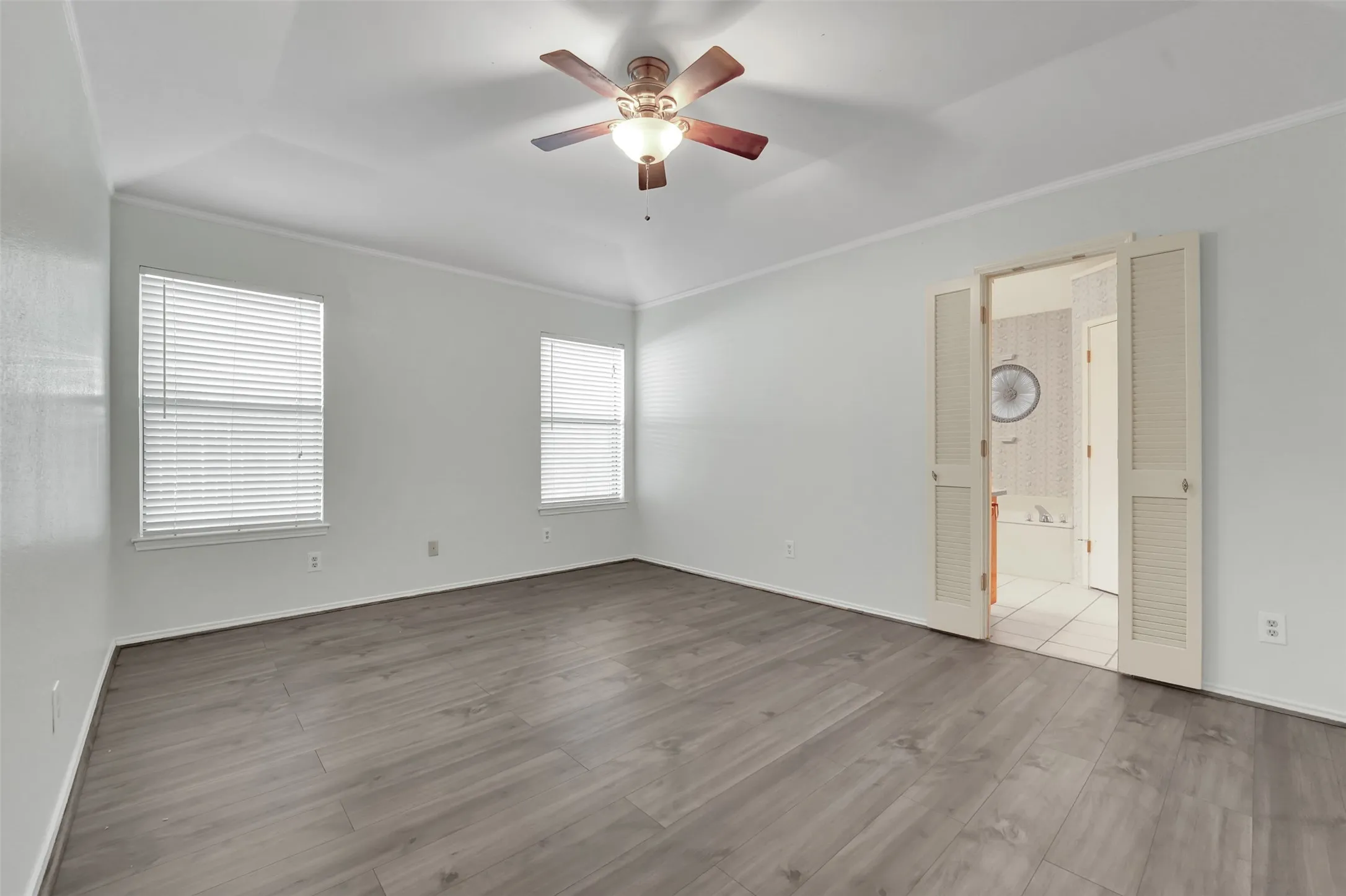 Vaulted ceiling with a ceiling fan, wood-finish flooring, two windows with blinds, and louvered double doors