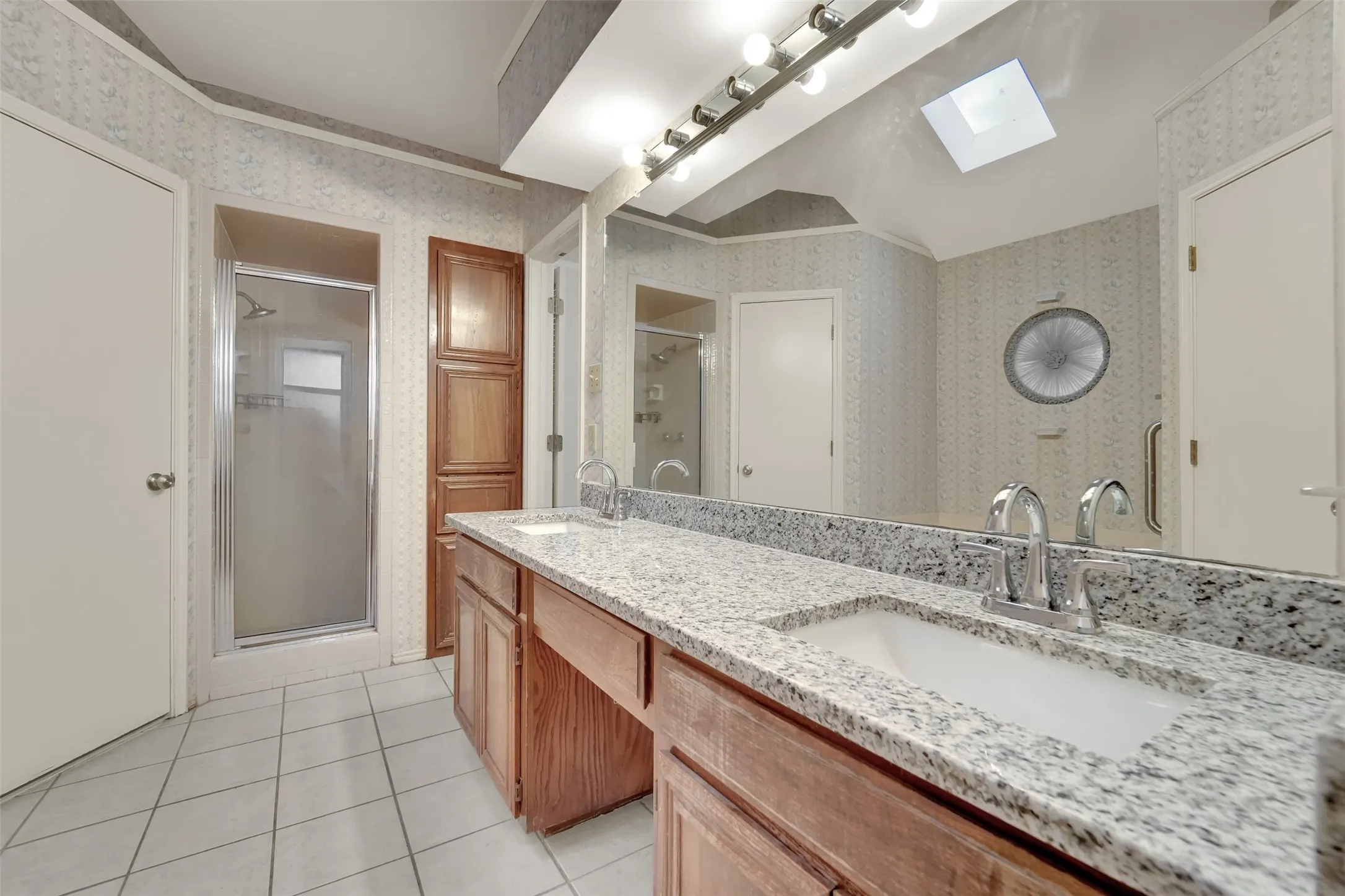 Dual vanity with granite countertops, wood-finish cabinetry, tile flooring, and a skylight