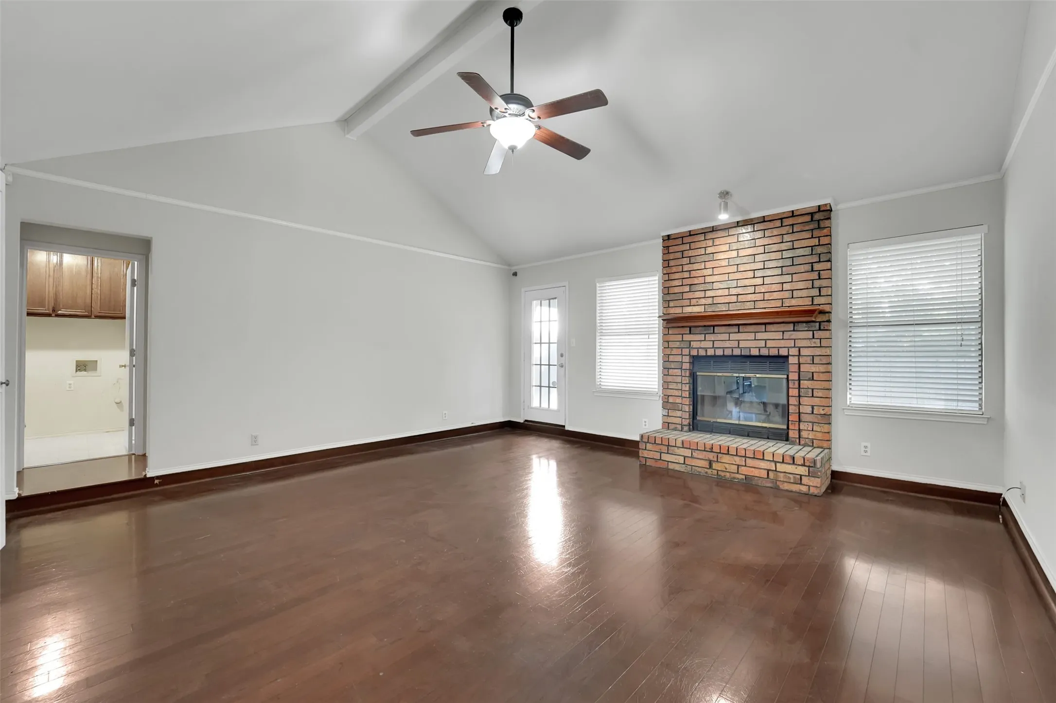 Vaulted ceiling with exposed beam, a ceiling fan, wood-finish flooring, brick fireplace with a wood mantle, and multiple windows with blinds