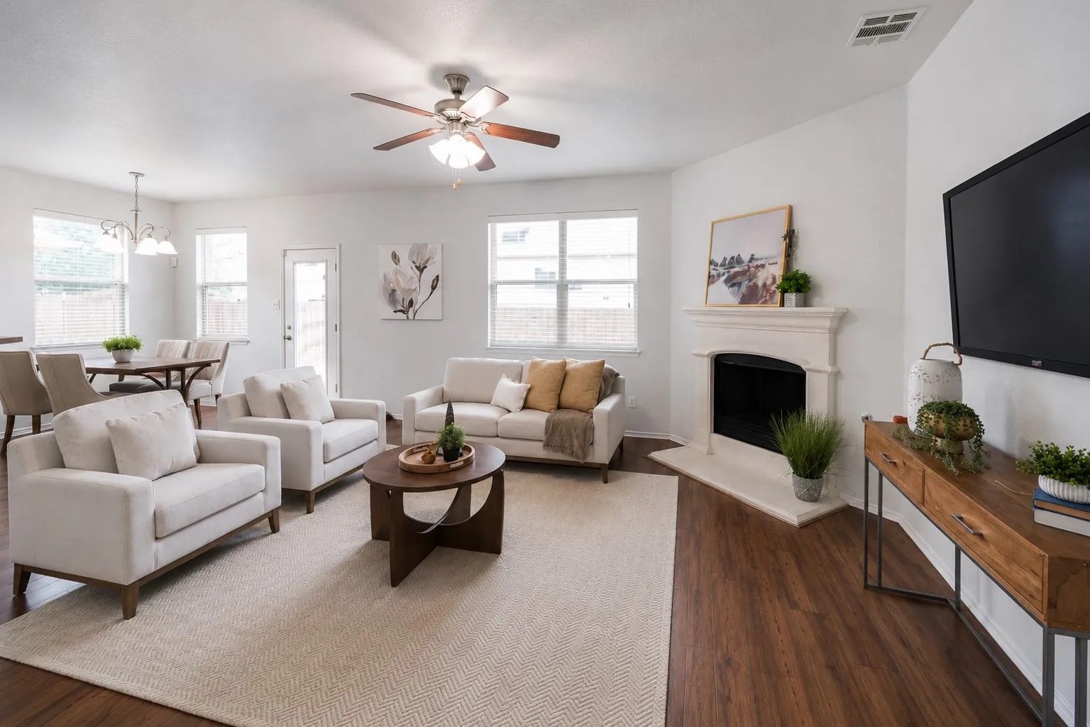 Expansive open-concept living area featuring wood-finish flooring, multiple windows, a ceiling fan with integrated lighting, a built-in fireplace with a mantel, and a dining area with a chandelier
