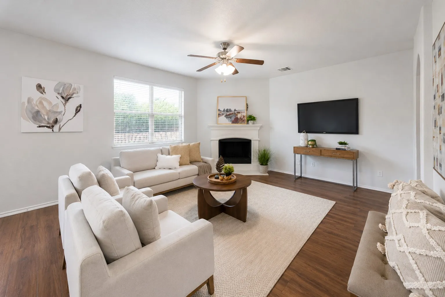 Living area featuring wood-finish flooring, a white fireplace with mantel, ceiling fan with light fixture, and multiple windows with blinds