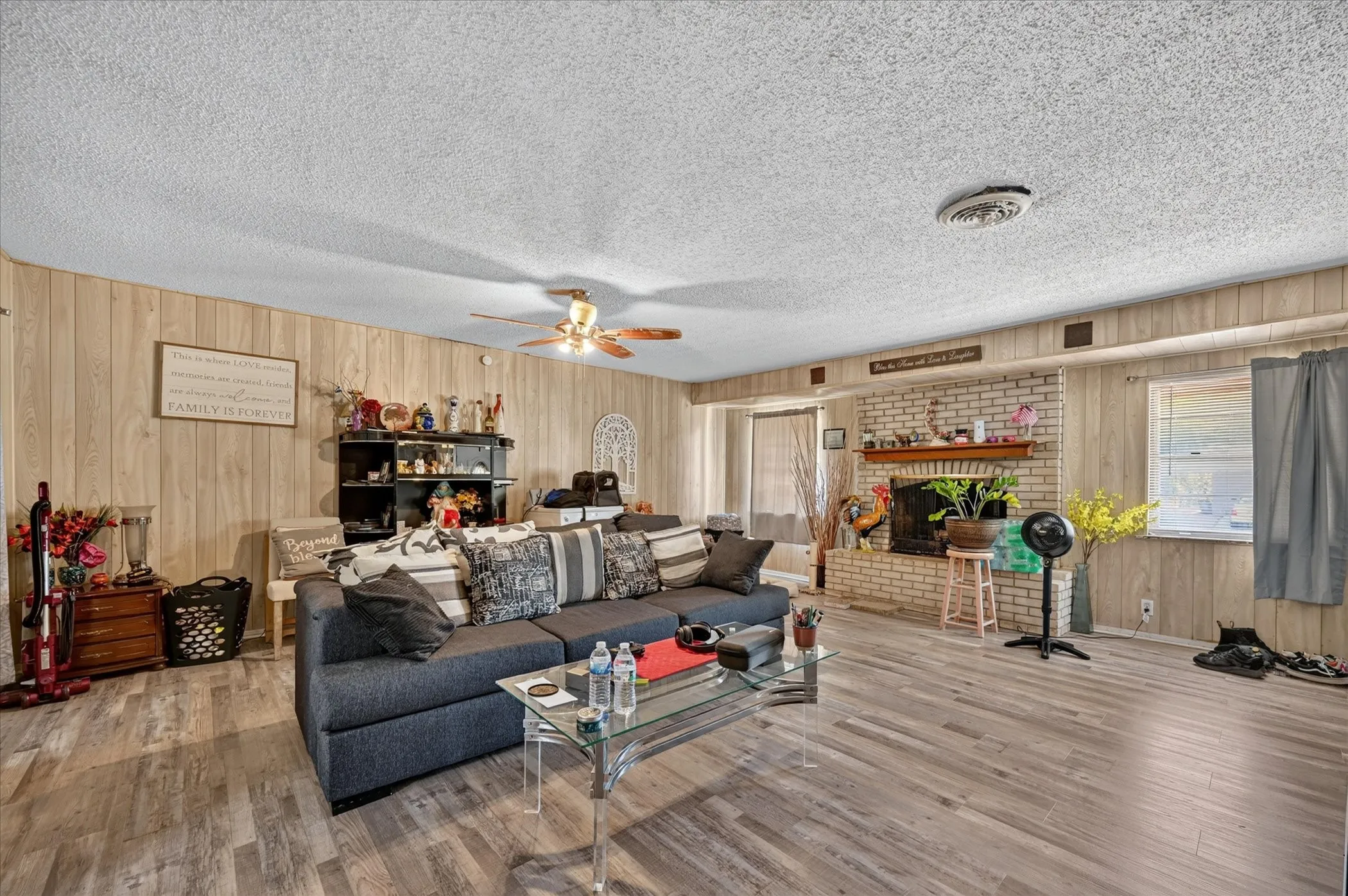 Expansive living area featuring wood-finish flooring, paneled wall accents, a brick-surround fireplace with a mantel, and an overhead ceiling fan