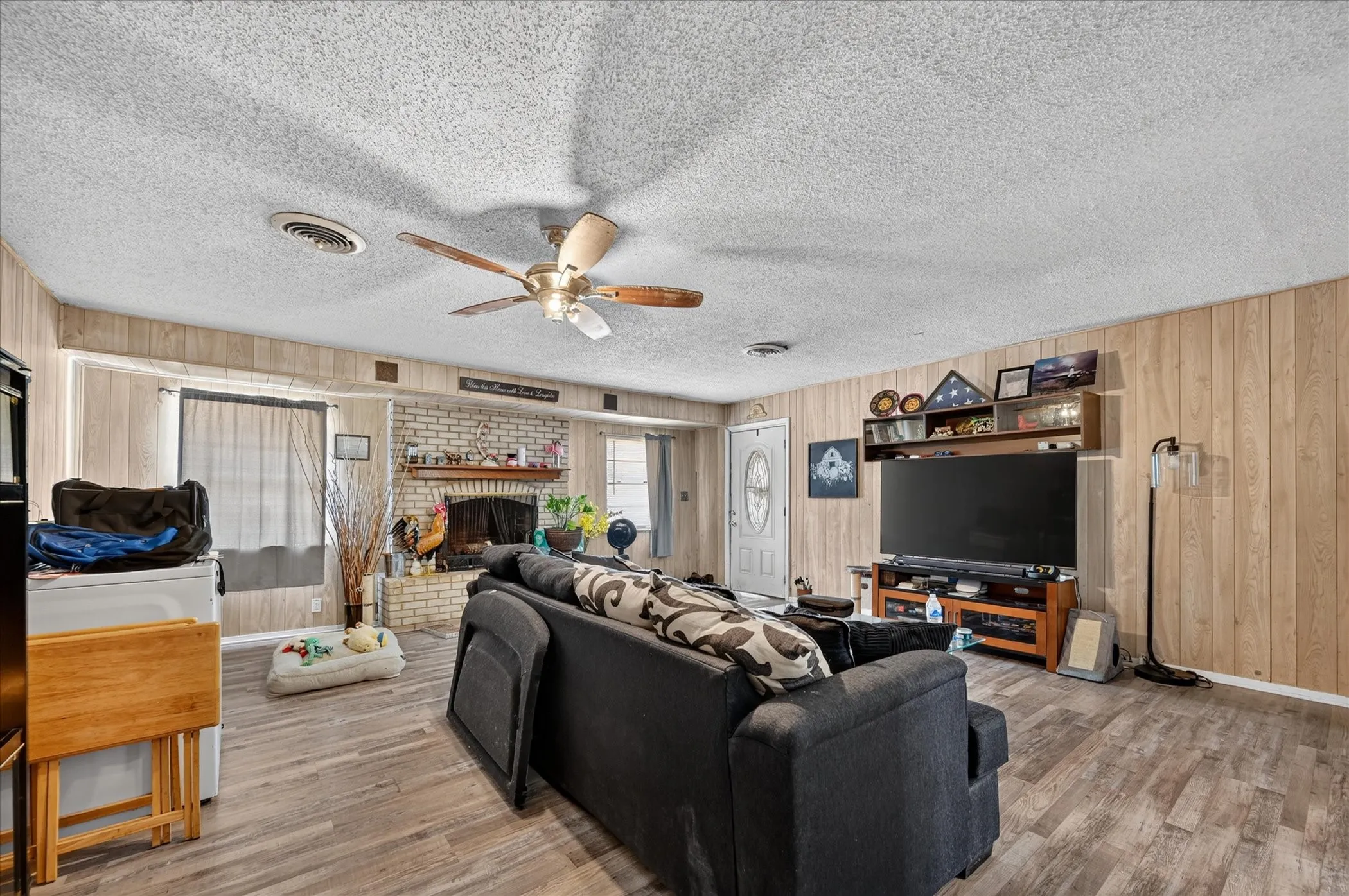 Living area featuring wood-finish flooring, paneled walls, a ceiling fan, and an exposed brick fireplace