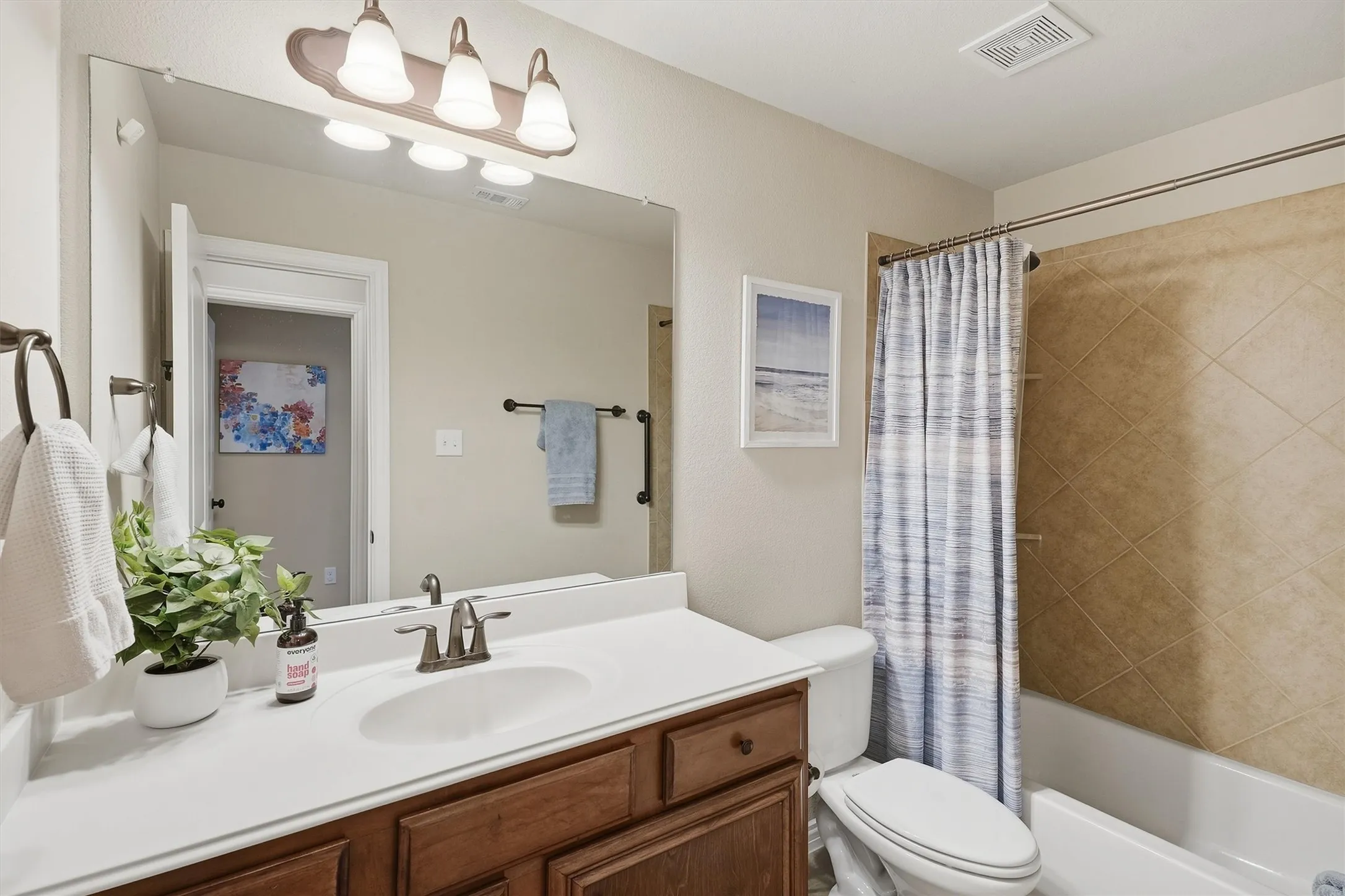 The secondary bathroom offers a single-sink vanity with warm wood cabinets, bronze fixtures, and a tub-and-shower combination featuring a tiled surround. Neutral paint and a large vanity mirror make it a bright, well-appointed space for family or guests.