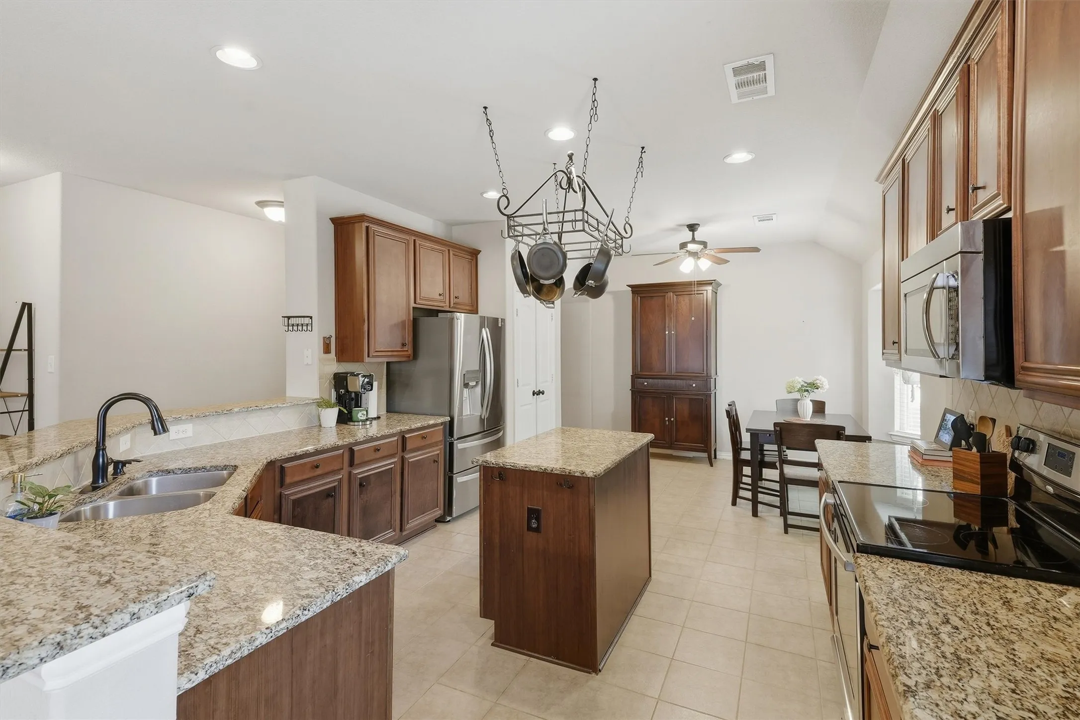 The spacious kitchen features rich wood cabinetry, durable granite countertops, stainless steel appliances, and a center island for extra prep space. An overhead hanging pot rack, breakfast bar, and seamless flow into the dining area make this setup ideal for meal prep and entertaining.