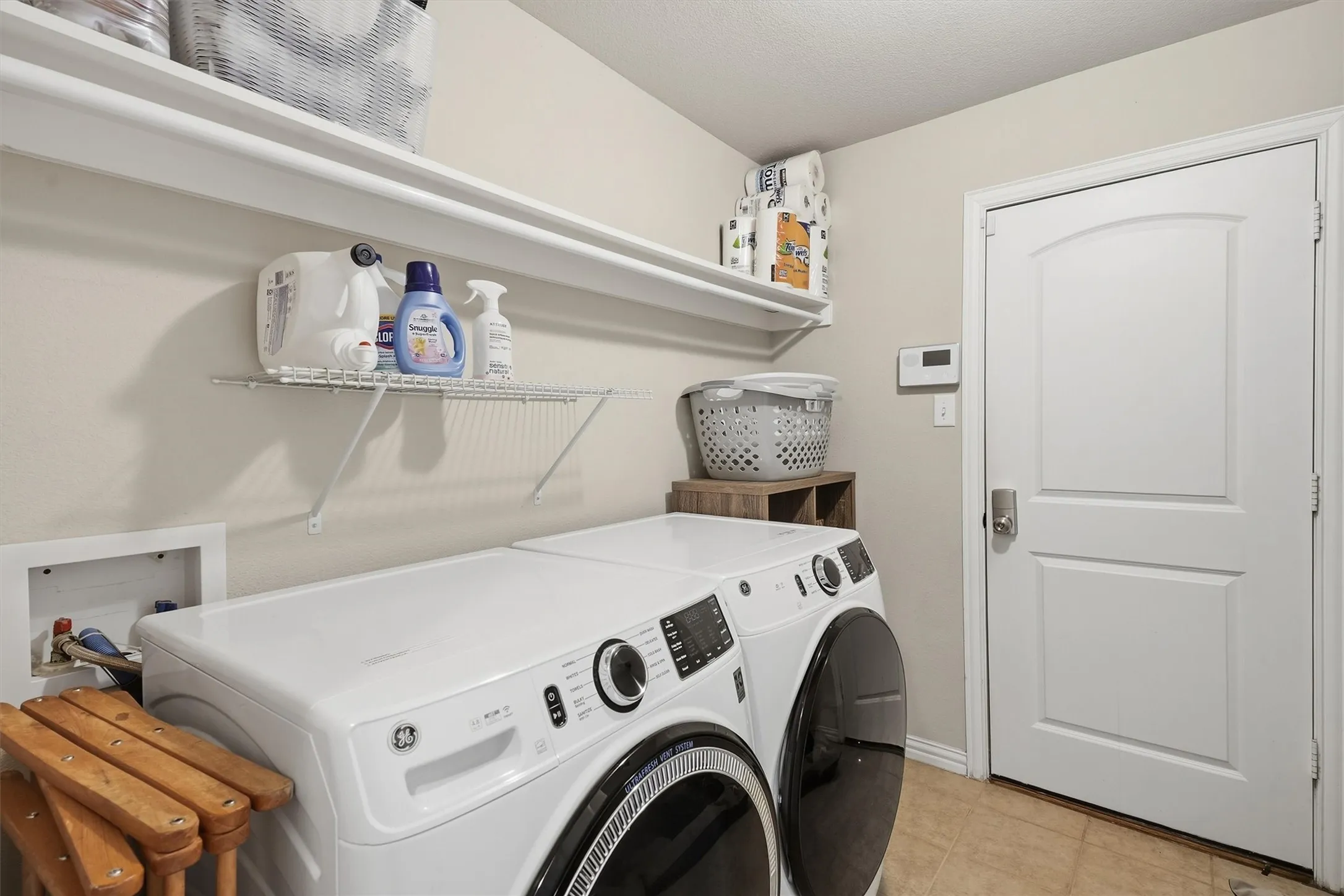 Convenient dedicated laundry room featuring tile flooring, built-in overhead wire shelving for detergent storage, and direct exterior door access.